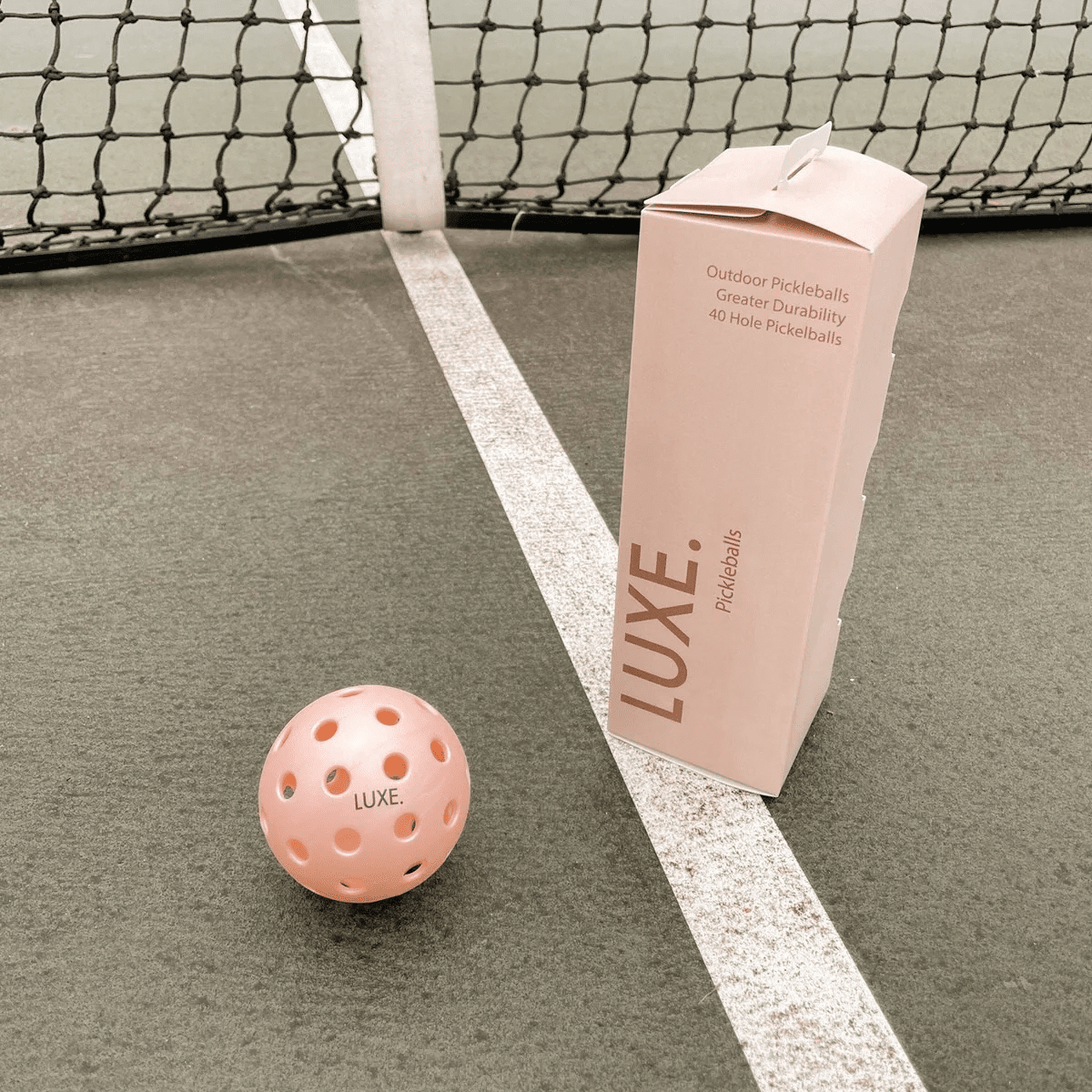A pink, high-visibility pickleball labeled “LUXE” sits near the court line. Next to it, a box displays "LUXE Pickleballs - Durable Practice and Tournament Balls for Indoor and Outdoor Play" by LUXE Pickleball. A tennis net stands behind.