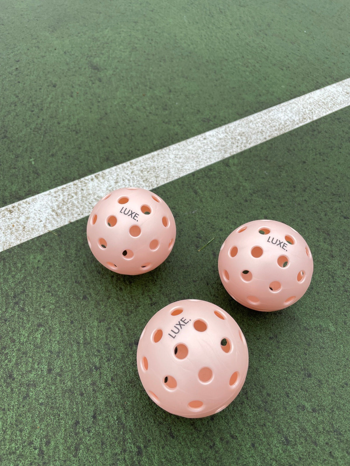 Three peach LUXE Pickleball balls, designed for indoor and outdoor play, rest on a green court near the white line. Their textured surfaces show signs of frequent use. Perfect for both practice and tournaments by LUXE Pickleball.