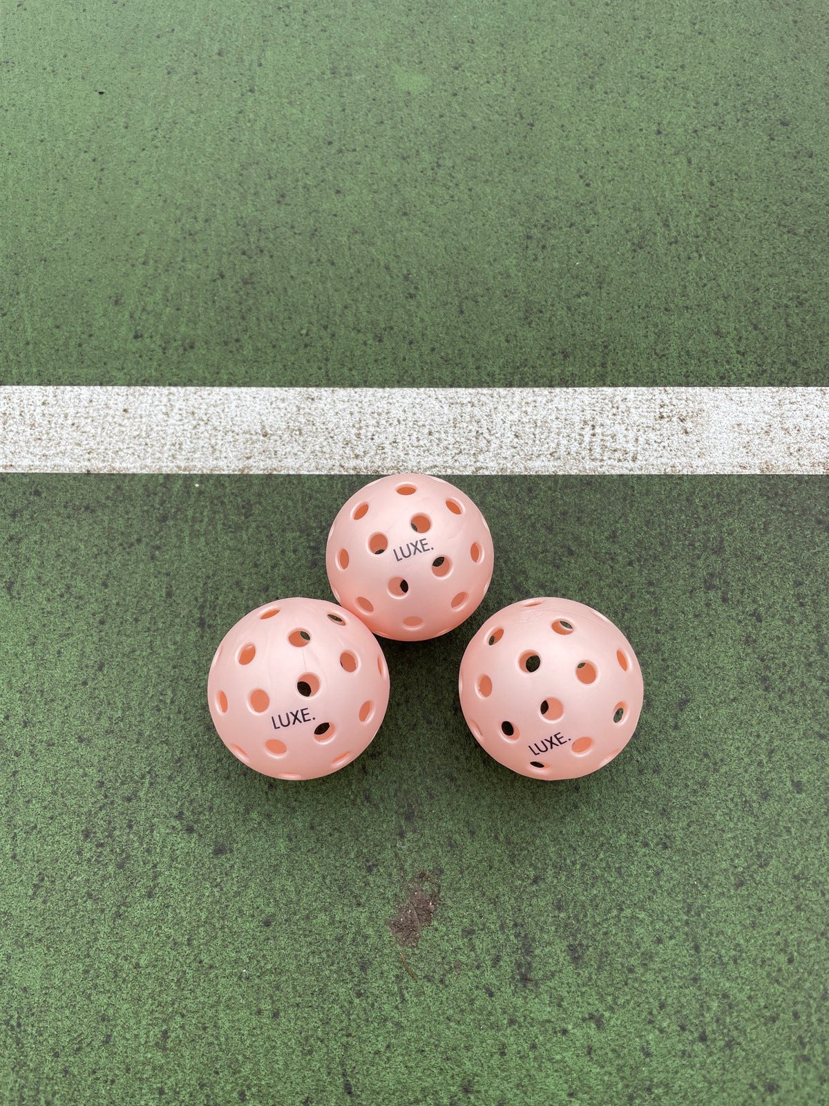 Three peach LUXE Pickleballs by LUXE Pickleball, designed for indoor and outdoor play, are arranged in a triangle on a green court with a wide white line in the background.
