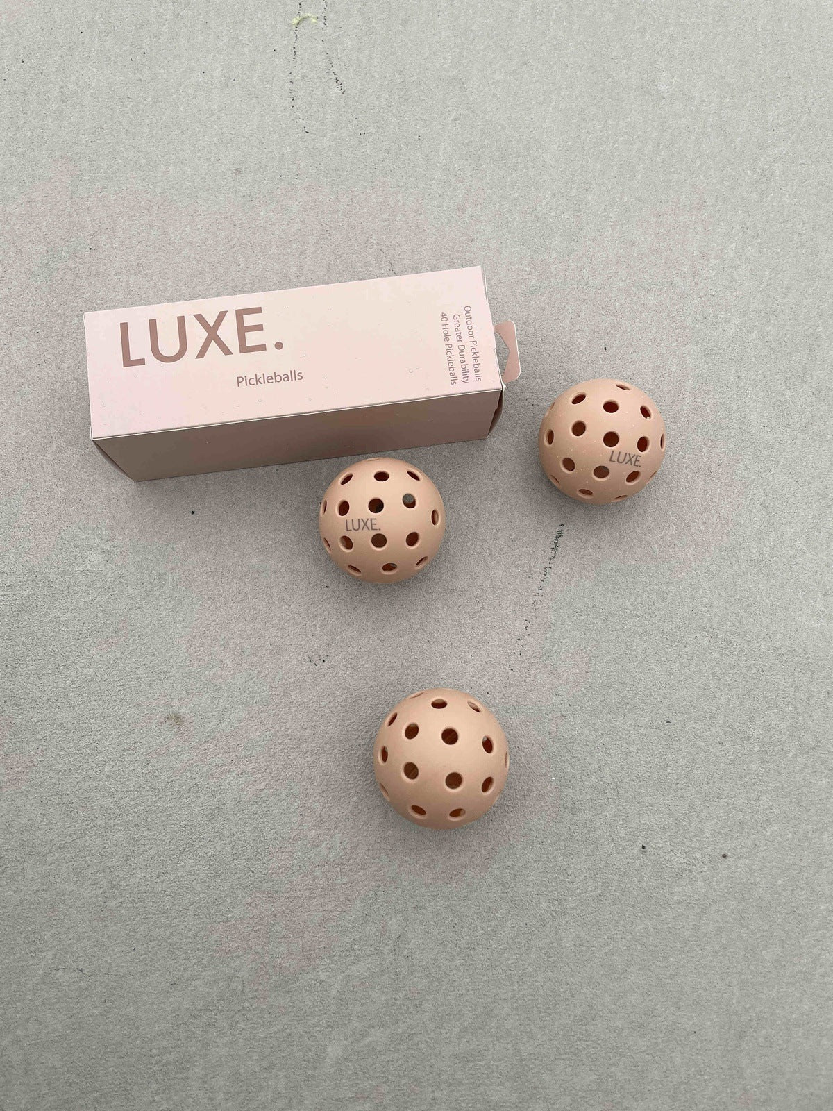 A rectangular pink box labeled “LUXE Pickleball” rests on a light grey surface, with three beige LUXE Pickleballs—durable for indoor/outdoor play—placed in front. Each ball features multiple holes and the brand name for a minimal, neutral look.