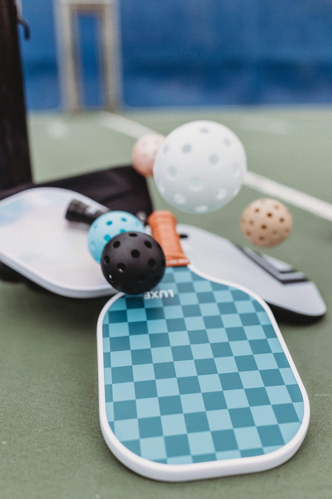 A LUXE Pickleball paddle rests on a green court with LUXE Pickleballs—durable, high-visibility practice and tournament balls in white, black, blue, and tan. A paddle case and net from LUXE Pickleball appear softly blurred in the background.