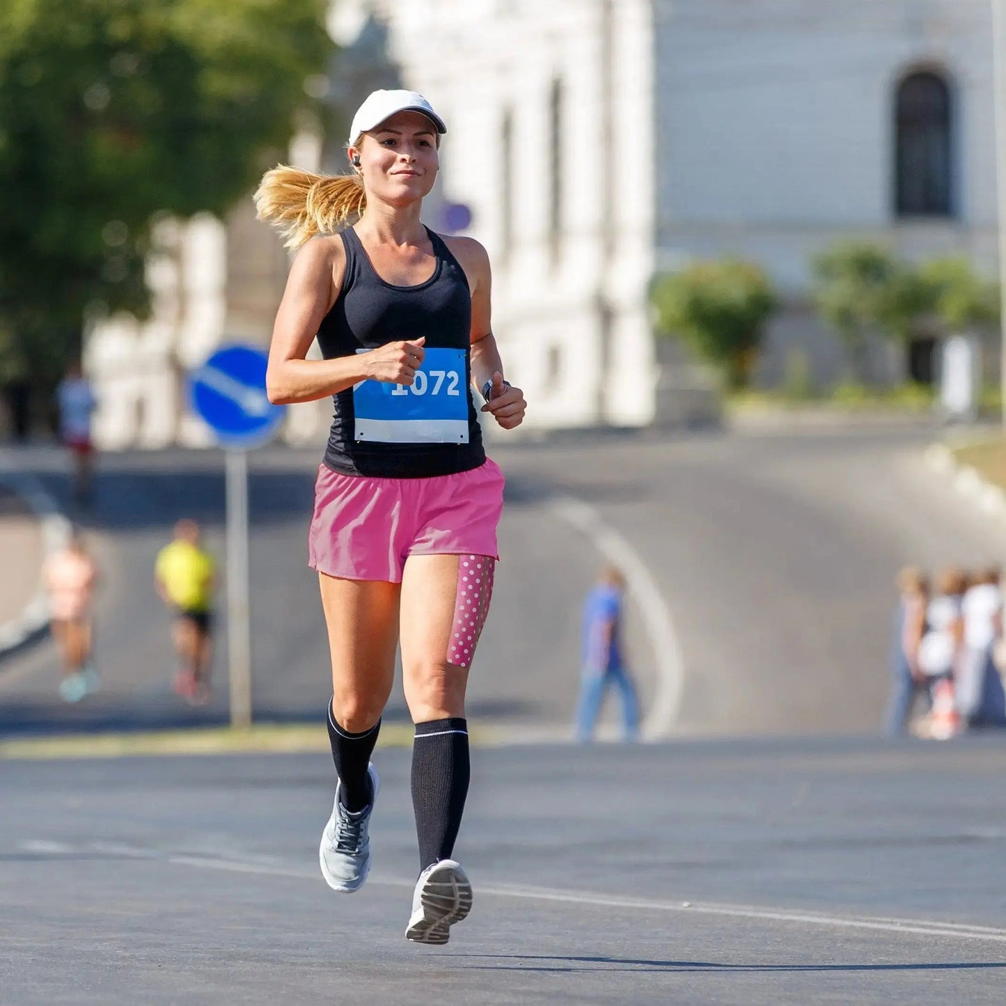 A woman races through a city street wearing KT Tape Pro® by KT Tape for athletic support, along with a white cap, black tank top, pink shorts, black compression socks, and a race bib. Blurred runners and spectators fill the tree-lined background.