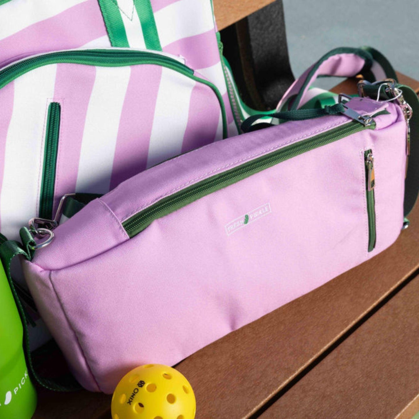 A light purple Fresh Pickle Insulated Cooler Bag with green zippers—great for court essentials—rests on a wooden bench. Behind it is a matching striped bag. A yellow pickleball and part of a green water bottle are also in view.