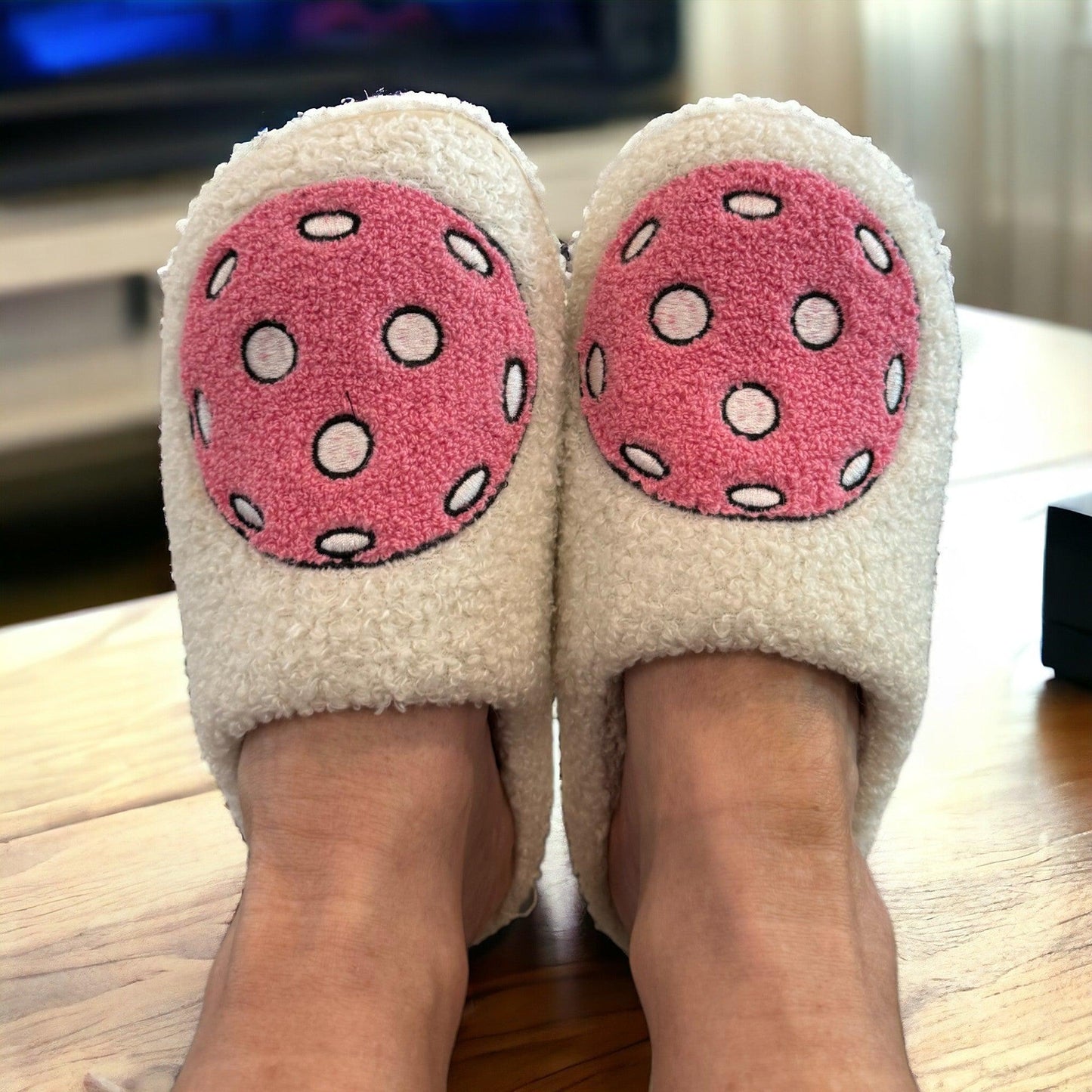 A pair of Fresh Pickle’s Pickleball Slippers, cozy in fuzzy white with playful pink circles and polka dots, are worn on relaxed feet atop a wooden surface. A blurred TV and gentle natural light enhance the comfy atmosphere.
