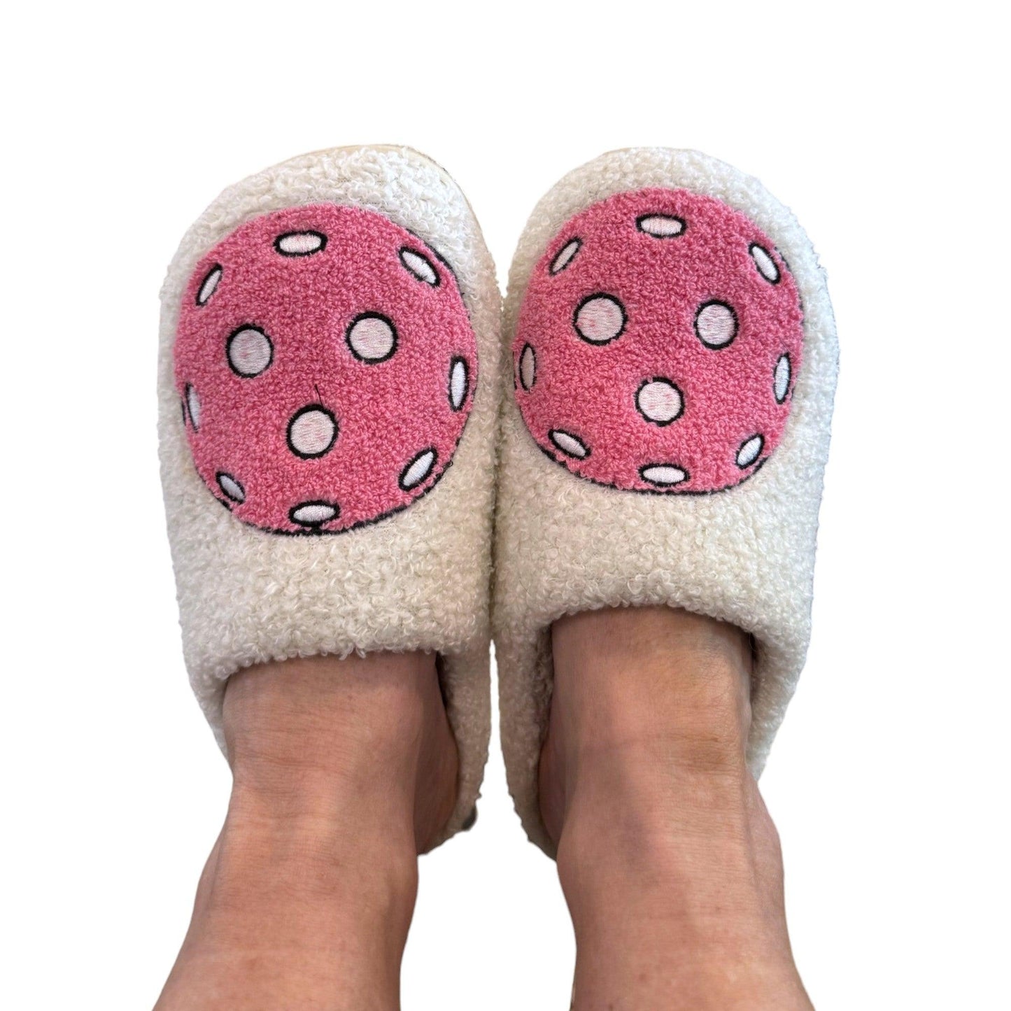 A pair of feet wears Fresh Pickle Pickleball Slippers, each cozy white slipper topped with a large pink circle patterned with white and black polka dots. The background is plain white.