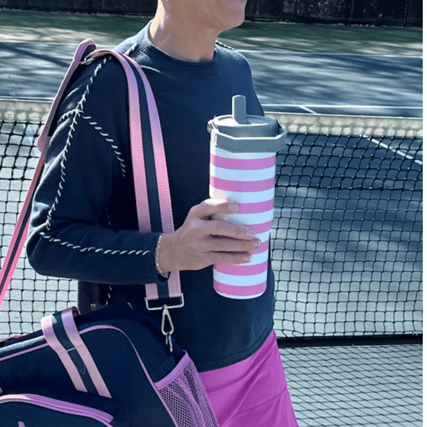 A person in a navy long-sleeve shirt and pink shorts carries a large navy gym bag with pink straps and holds the Fresh Pickle 30 oz double-walled vacuum sealed Fresh Pickle Tumbler with flip-up straw. A tennis court with black mesh fence is in the background.