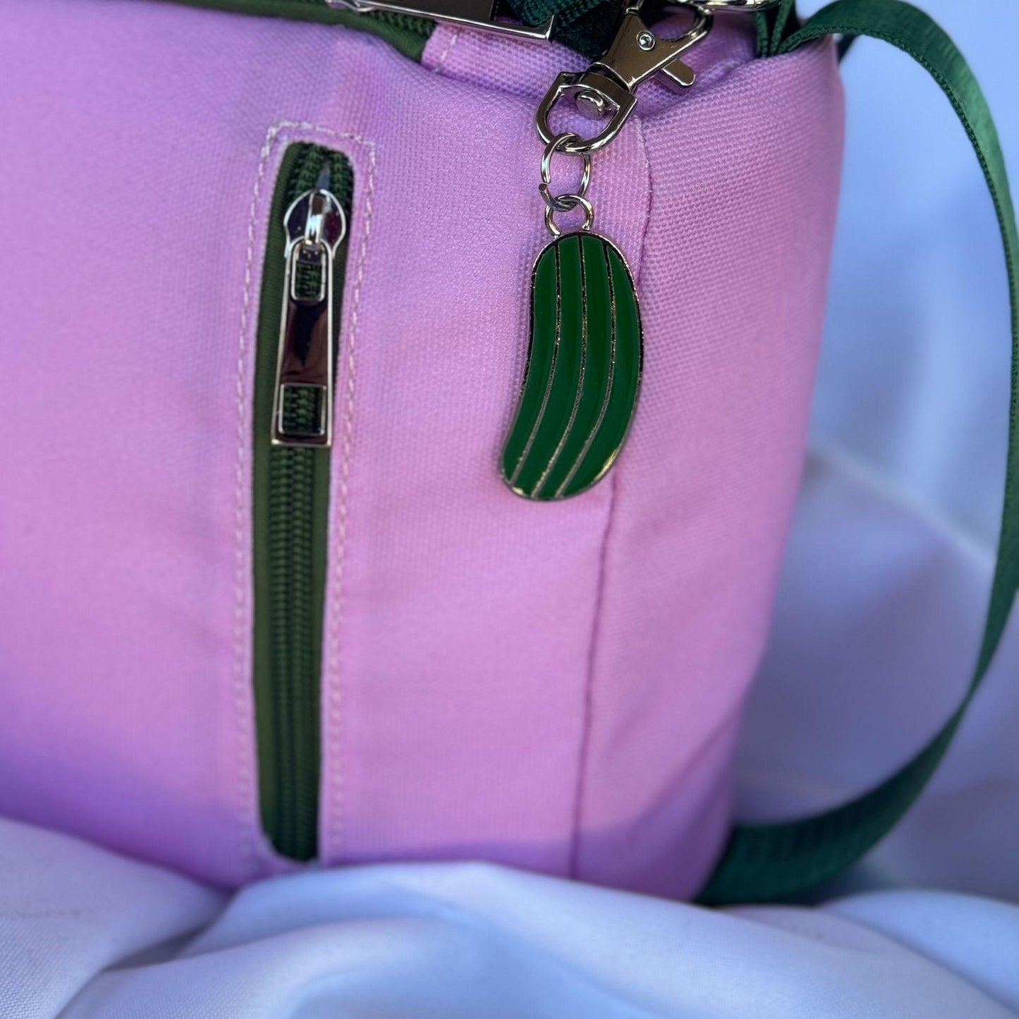 A close-up of the Fresh Pickle Insulated Cooler Bag in pink with a green zipper and cucumber-shaped keychain. The bag rests on white fabric, and a green strap for court essentials is visible at the edge.