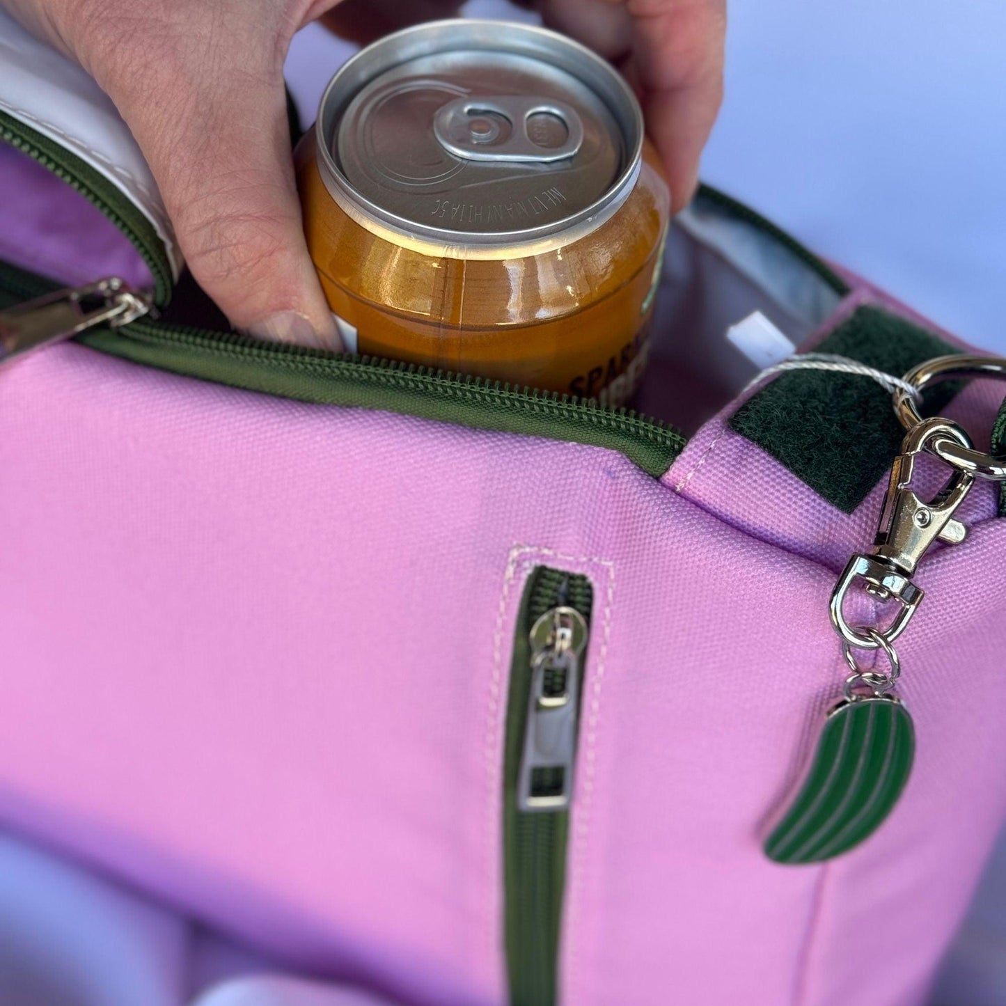 A hand places a can of sparkling water into the Fresh Pickle Insulated Cooler Bag, which is pink with green accents, a silver zipper, outer pocket, and cactus-shaped keychain. The background is softly blurred.