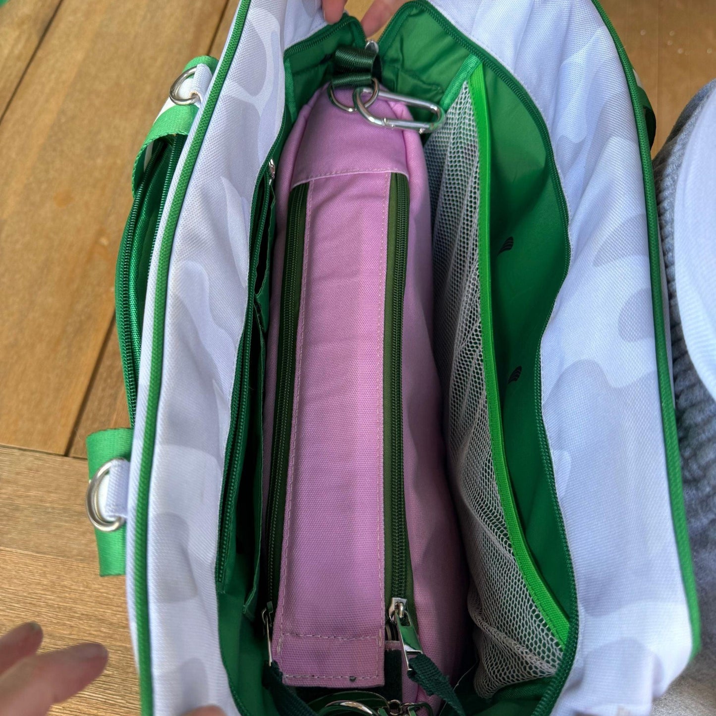 A top-down view of the Fresh Pickle Insulated Cooler Bag in green and white camo. Inside, a pink rectangular pouch with a green zipper and mesh pockets stores essentials. Hands grip the soft-sided cooler on a wooden surface.