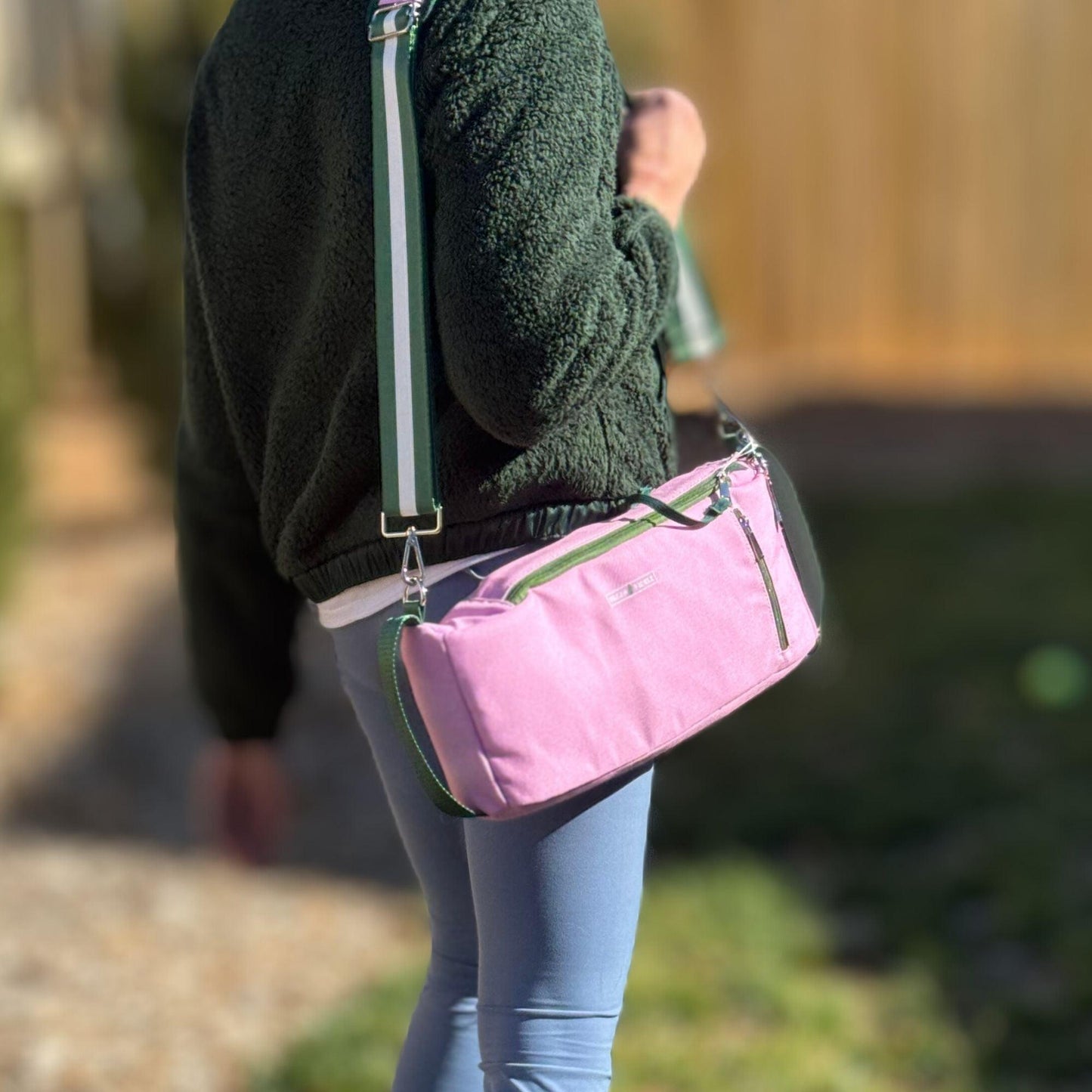 A person in a dark green fuzzy jacket and light blue leggings carries the Fresh Pickle Insulated Cooler Bag, which is pink with green accents, silver zippers, and a white and green strap. Grass and a wooden fence are visible in the background.