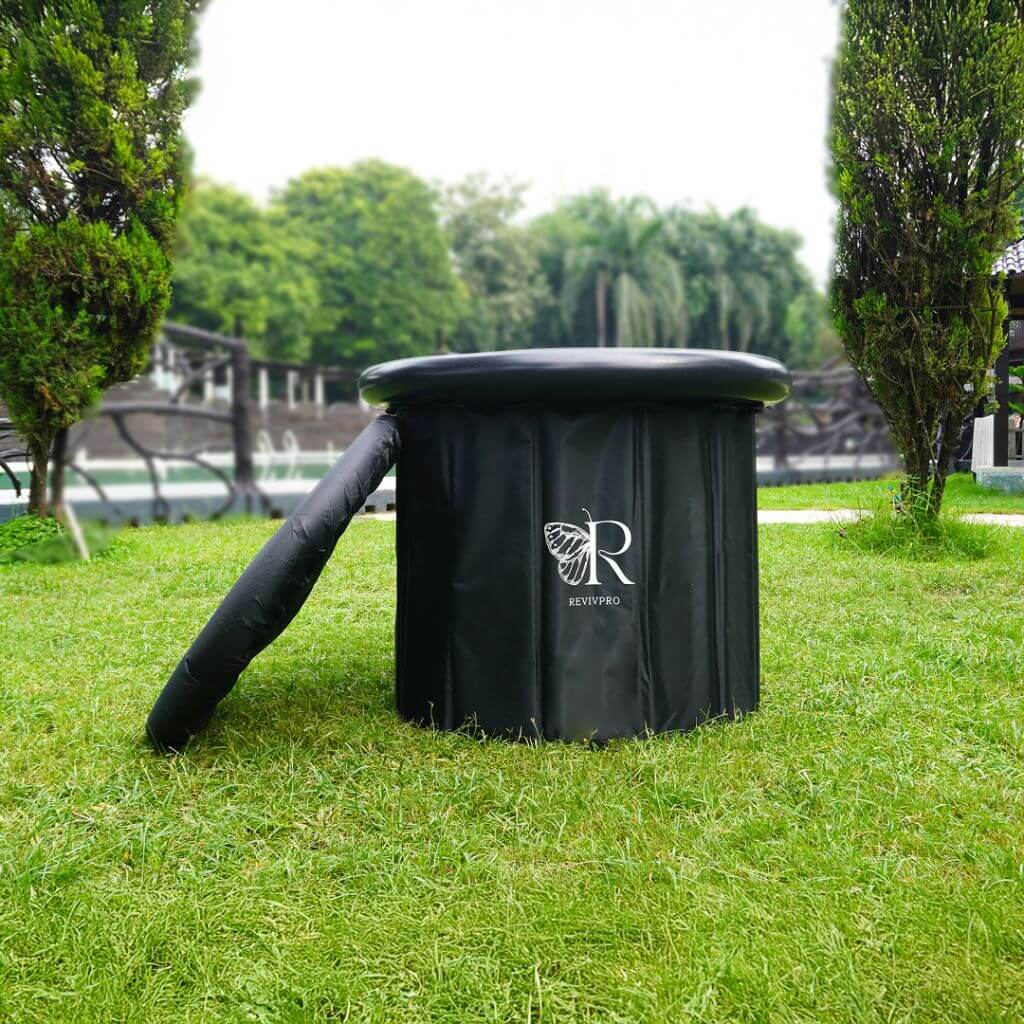 A black, cylindrical RevivPro Portable Foldable Ice Bath Tub—water chiller compatible for recovery and wellness—sits on grass between two trimmed bushes. The white RevivPro logo is visible; trees and a fence are in the background under a clear sky.
