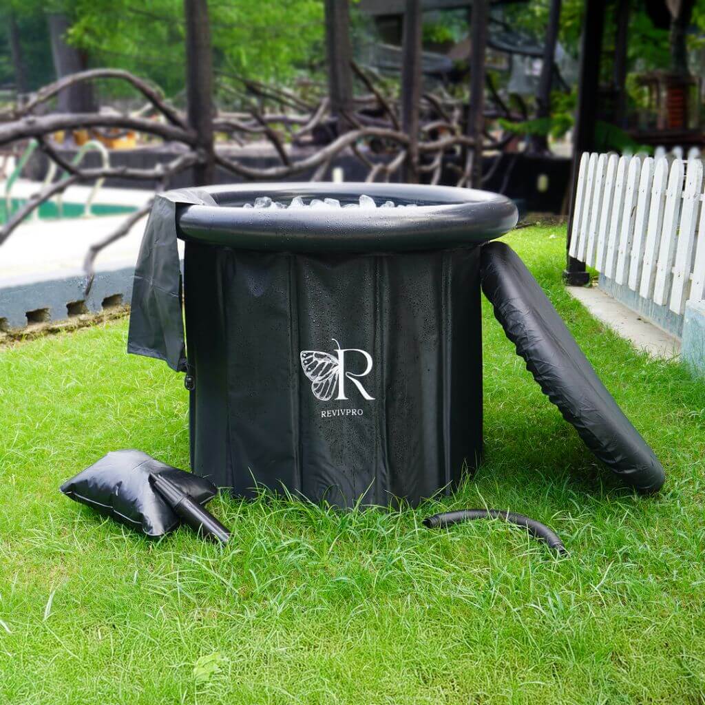 A round, black RevivPro Portable Foldable Ice Bath Tub sits on grass, filled with water for cold plunge therapy. Nearby are a cover, pillow, and towel; a white picket fence and lush trees enhance the wellness scene.