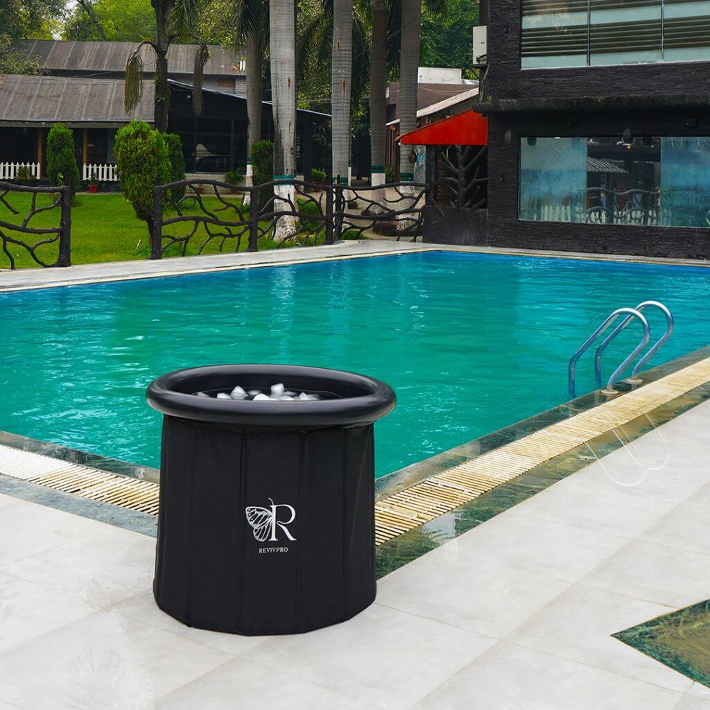 A RevivPro Portable Foldable Ice Bath Tub, water chiller compatible and branded with an “R” and leaf logo, sits filled with ice by a blue pool, offering outdoor cold plunge therapy among palm trees, grass, and modern buildings.