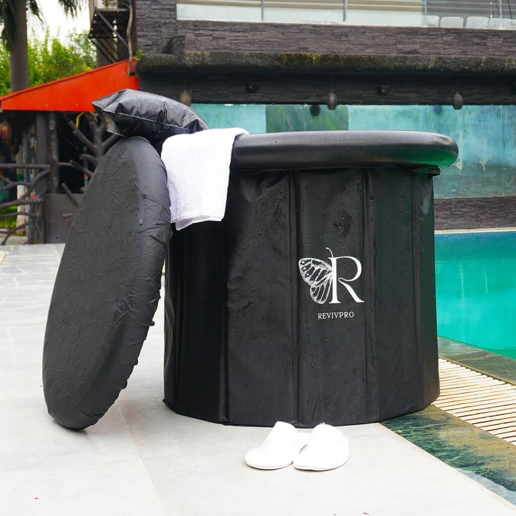 A black RevivPro Portable Foldable Ice Bath Tub sits poolside, lid propped nearby with a white towel and headrest on top. White slippers rest close. Enjoy cold plunge recovery therapy with views of the pool, greenery, and a modern building backdrop.