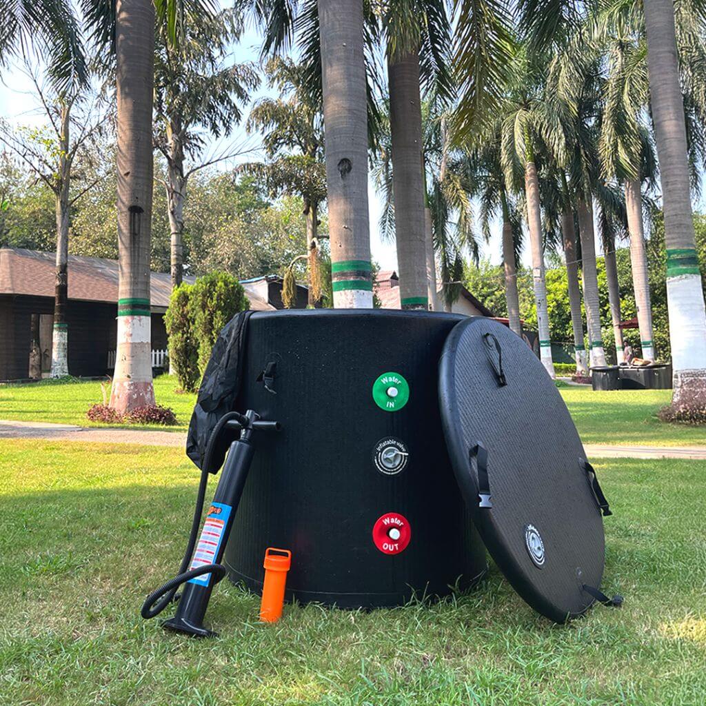 The RevivPro Portable Ice Bath Tub Pro, a water chiller-compatible cold plunge recovery solution with IN/OUT valve buttons, pump, and whistle sits on green grass near its lid, surrounded by palm trees and a house under the sunny sky.