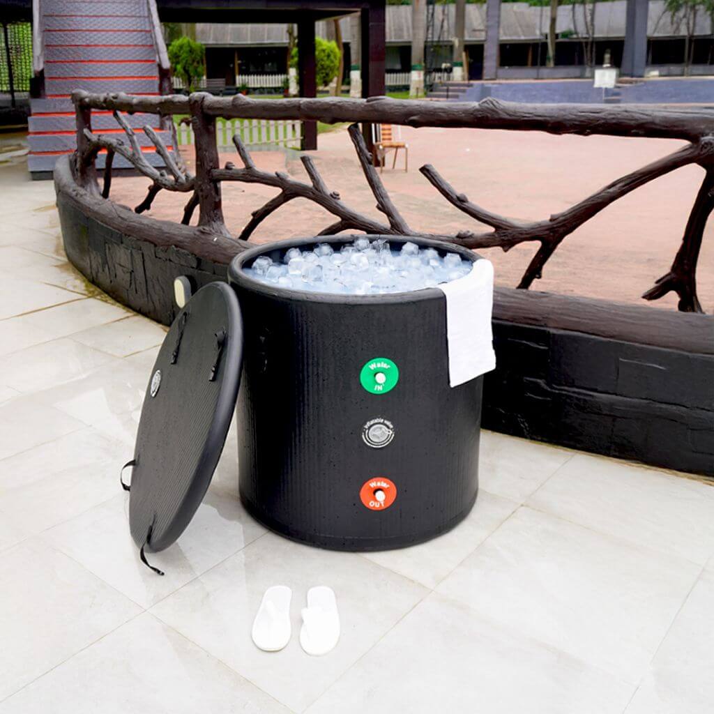The RevivPro Portable Ice Bath Tub Pro, a water chiller compatible cold plunge recovery solution, sits on a tiled patio with ice cubes and a white towel draped over the rim; black lid and white sandals rest nearby by a rustic wooden railing.