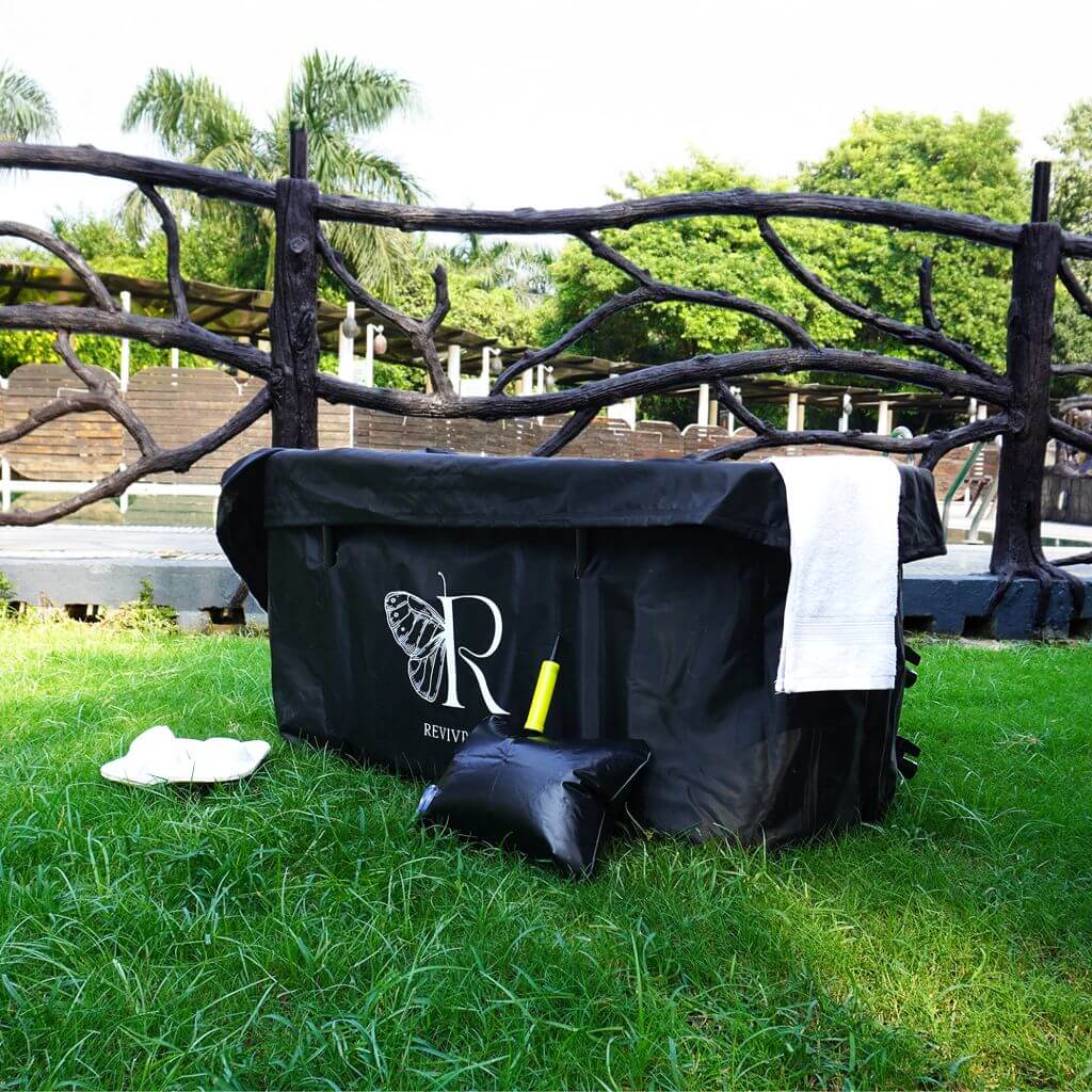 The RevivPro Portable Oval Ice Bath Tub X with Extra Length stands on green grass, featuring a black foldable design with an "R" and butterfly logo. A white towel rests on its edge, with a white cloth, black pillow, and yellow item nearby.