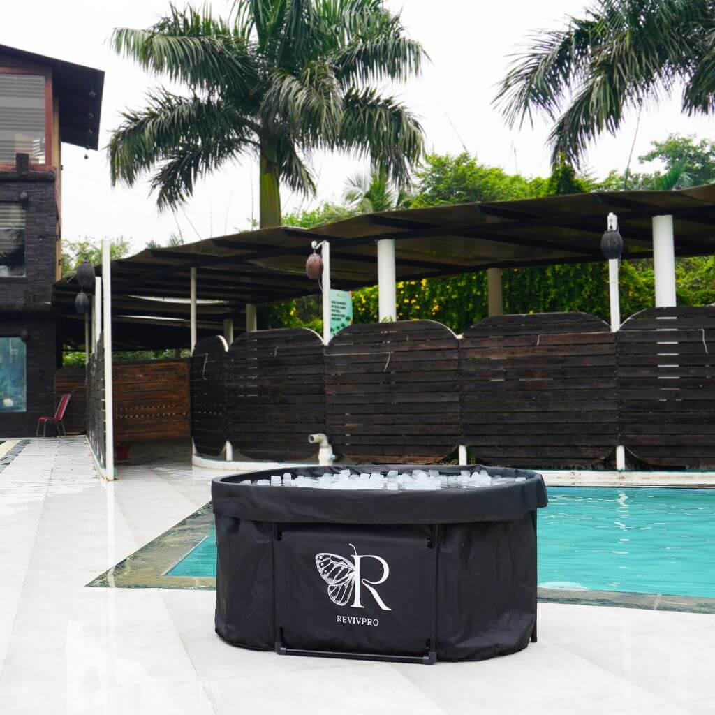 A RevivPro Portable Oval Ice Bath Tub X with extra length, water chiller compatible, sits on white tiles by a pool amid palm trees and wooden privacy barriers, with a modern building in the background.