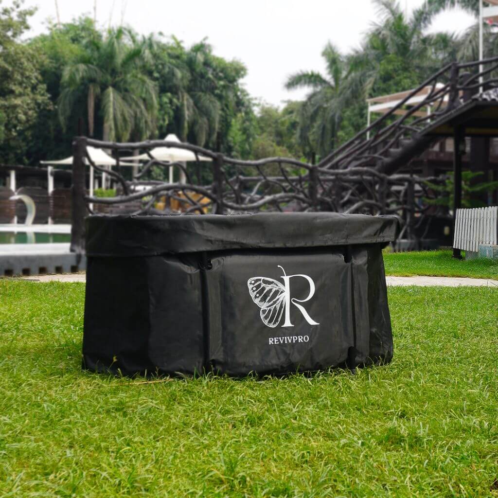 A black, oval RevivPro Portable Ice Bath Tub X with extra length, showing the butterfly R logo, is placed on green grass outdoors, surrounded by palm trees, a pool, and a wooden bridge in the background—water chiller compatible.