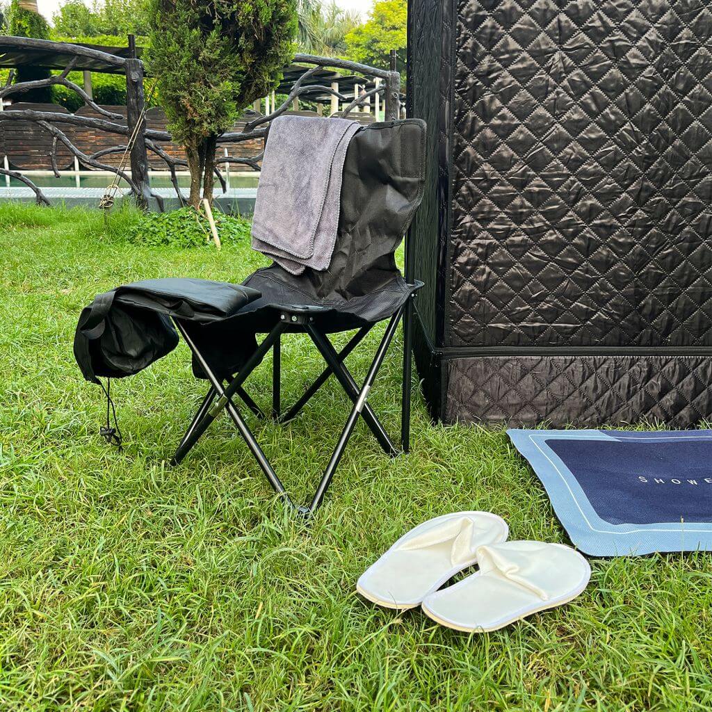 A black RevivPro Portable Steam Sauna Box with Steamer sits on the grass next to a folded black chair draped with a gray towel. Nearby, a navy blue mat holds white slippers. Trees and a wooden fence are in the background.