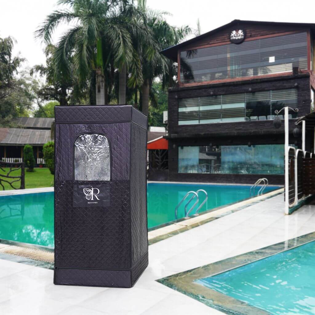 A black, textured RevivPro Portable Steam Sauna Box with Steamer featuring a clear window and white "R" logo stands on a wet poolside patio, backed by two pools, palm trees, and a modern building with large windows and outdoor seating.