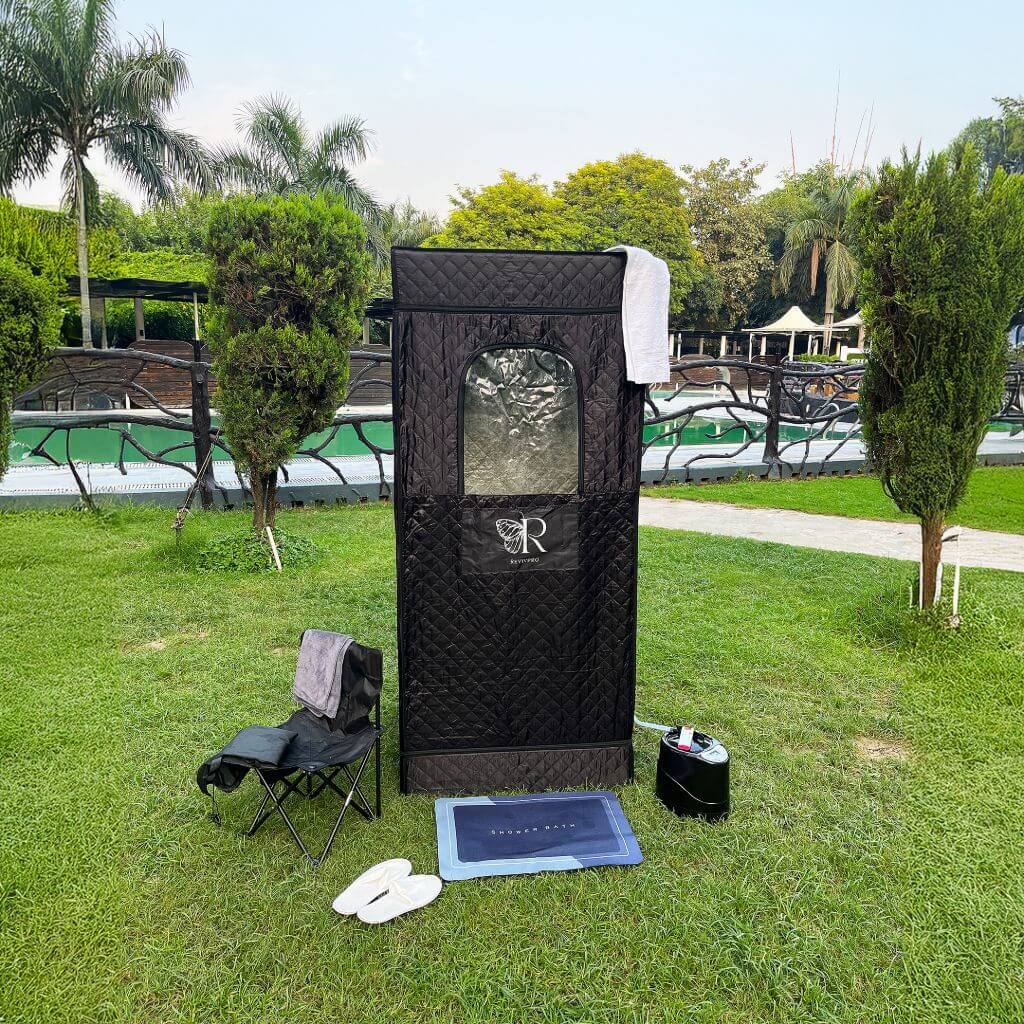 A RevivPro Portable Steam Sauna Box with Steamer stands on the grass like a home spa, topped with a towel. A folding chair, white slippers, mat, and black water heater sit in front, set against trees, a fence, and pool.