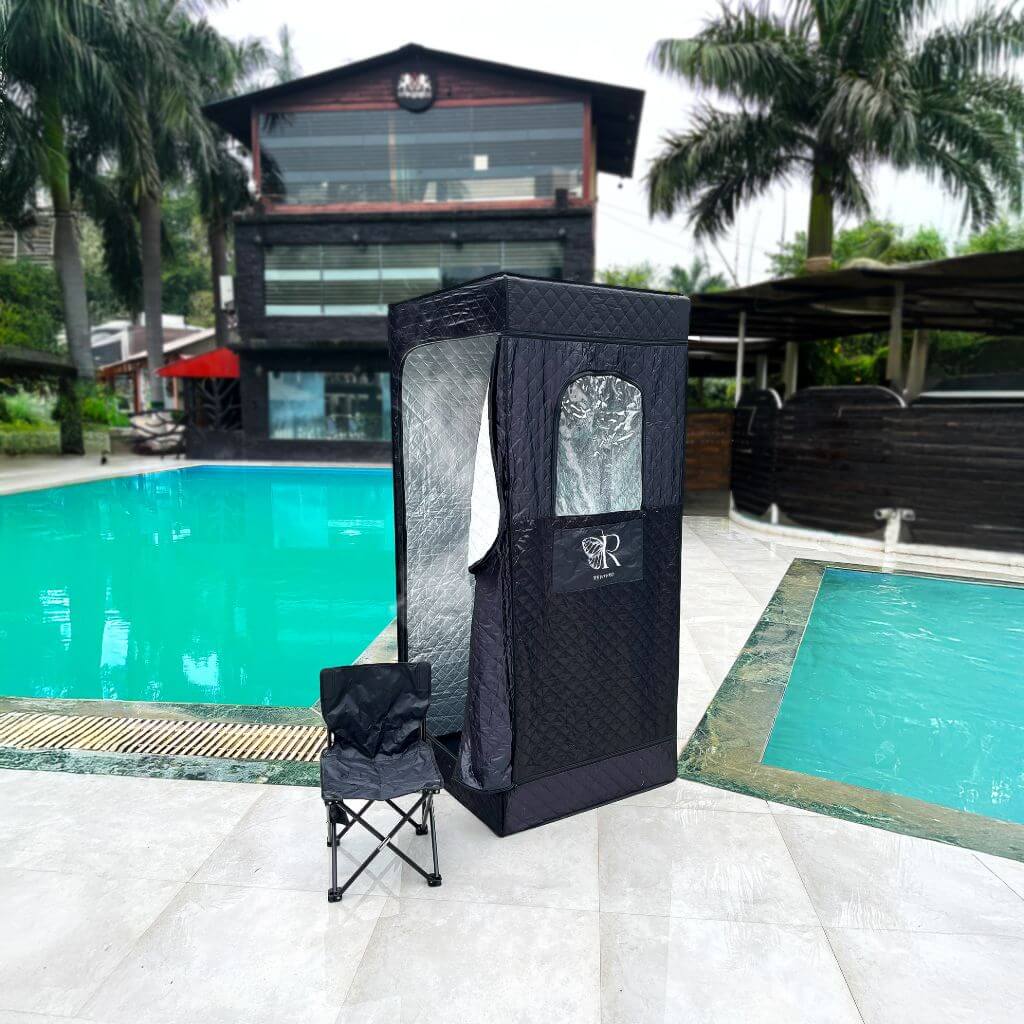 The RevivPro Portable Steam Sauna Box with Steamer stands by a pool next to a folded chair. Tropical palm trees and a modern multi-story building with large windows create a stylish backdrop.