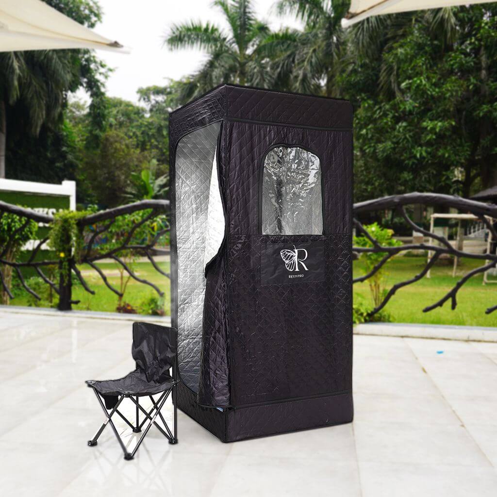 A black, rectangular RevivPro Portable Steam Sauna Box with Steamer, featuring a zippered door, clear window, quilted texture, and reflective interior, stands on a patio with a folded black camping chair nearby and lush greenery in the background.