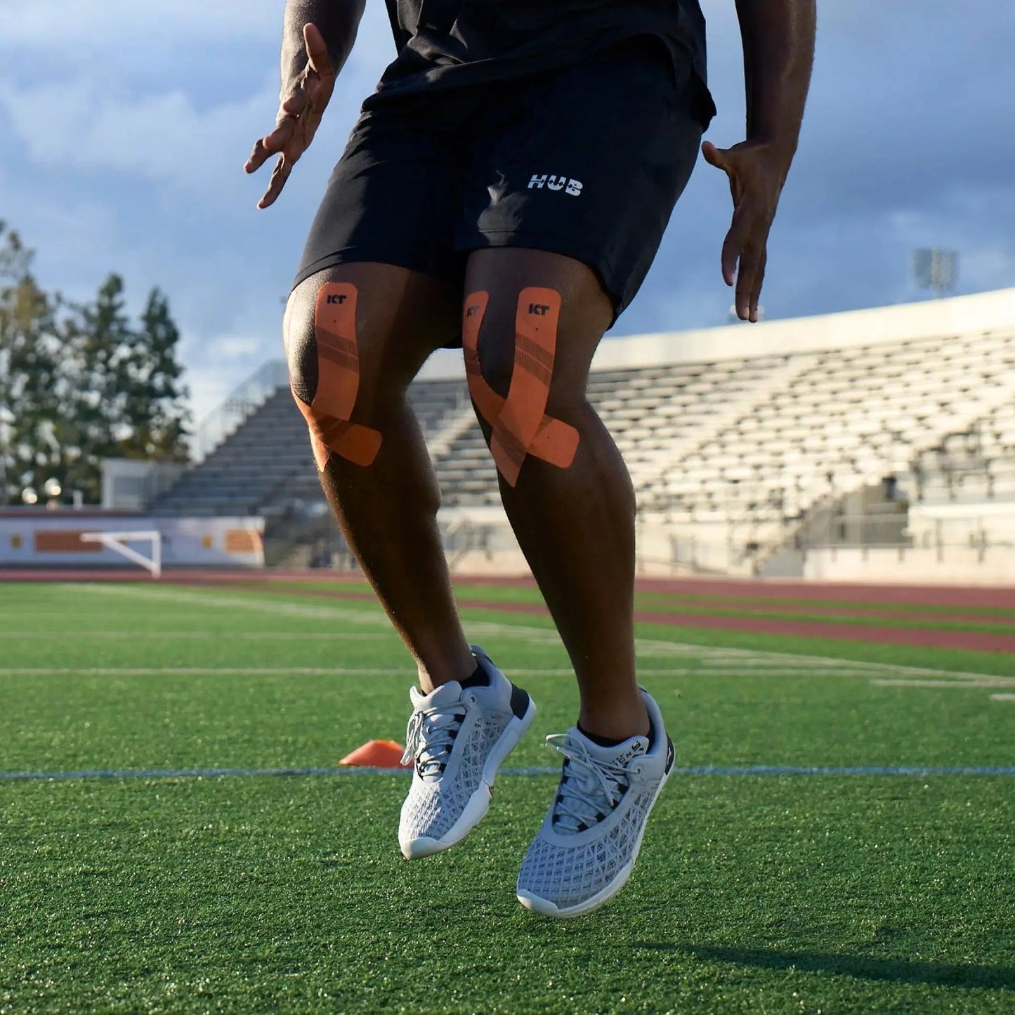 A person wearing black shorts and gray athletic shoes jumps on a green field, both knees supported by KT Tape Pro® from KT Tape. Only the lower body is visible; stadium seats and trees appear in the background under a clear sky.
