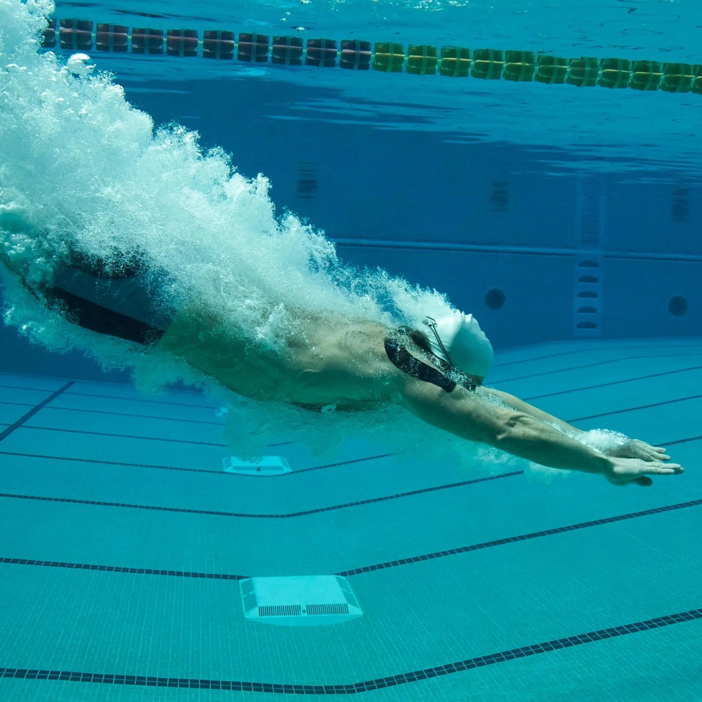 A swimmer in goggles and a cap glides underwater, arms extended in streamline form. Wearing KT Tape Pro Extreme® by KT Tape for pain relief, they leave bubbles behind as sunlight dances on the blue pool tiles.
