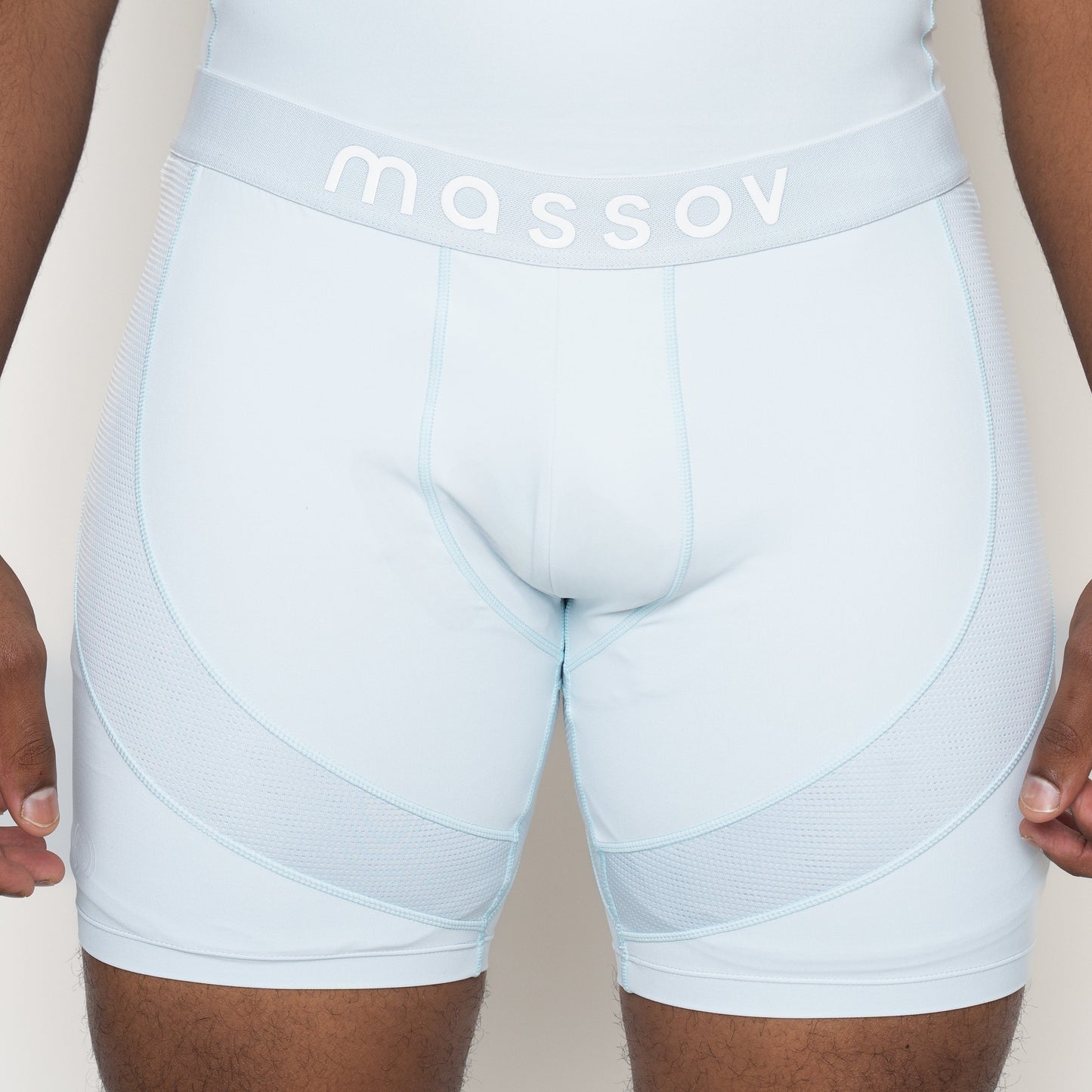 A close-up of a person wearing Massov ProForm® Boys' 7″ Compression Shorts in light blue, featuring mesh panels and a thick waistband with "massov" in white. The person's hands are relaxed by their sides.