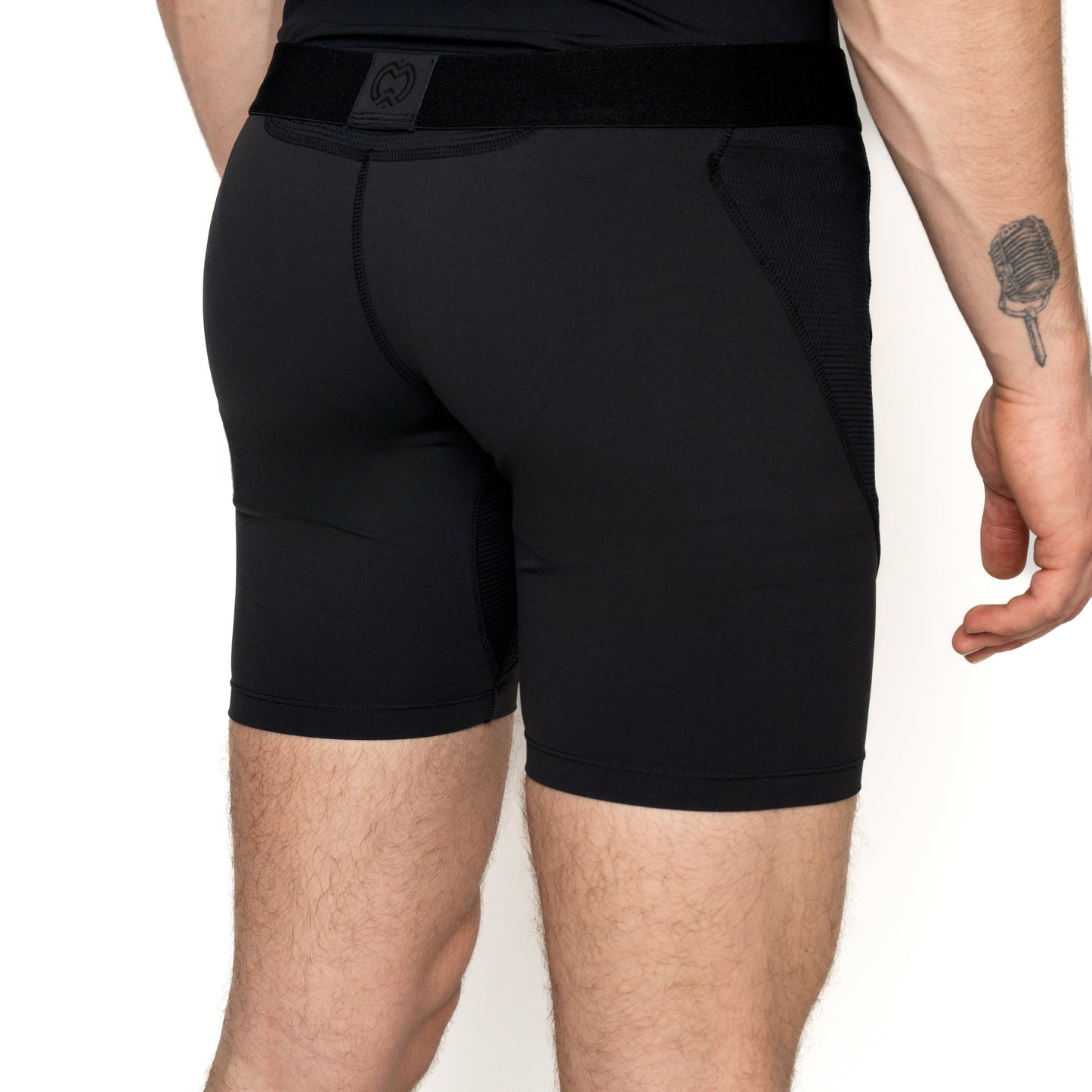 A person faces away, wearing Massov ProForm® Boys' 7″ Compression Shorts. Their left forearm displays a vintage microphone tattoo. The background is plain white.