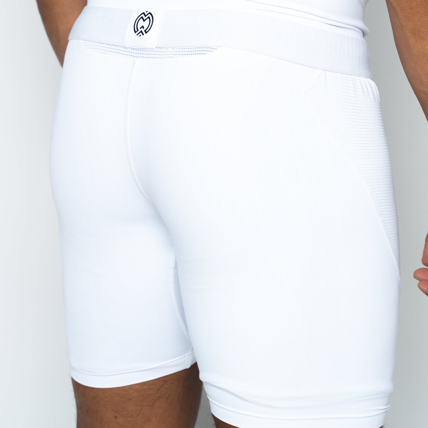 A close-up of Massov ProForm® Boys' 7″ Compression Shorts in white shows the back and side, emphasizing the smooth texture, seams, and elastic ProForm waistband with a small center logo, plus part of the boy’s arm and leg.