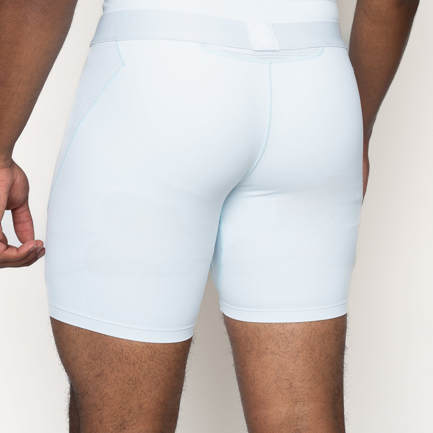 A youth boy is shown from the waist down in light blue Massov ProForm® Boys' 7″ Compression Shorts. The back view highlights the smooth ProForm fabric and close fit, with one arm visible against an off-white background.