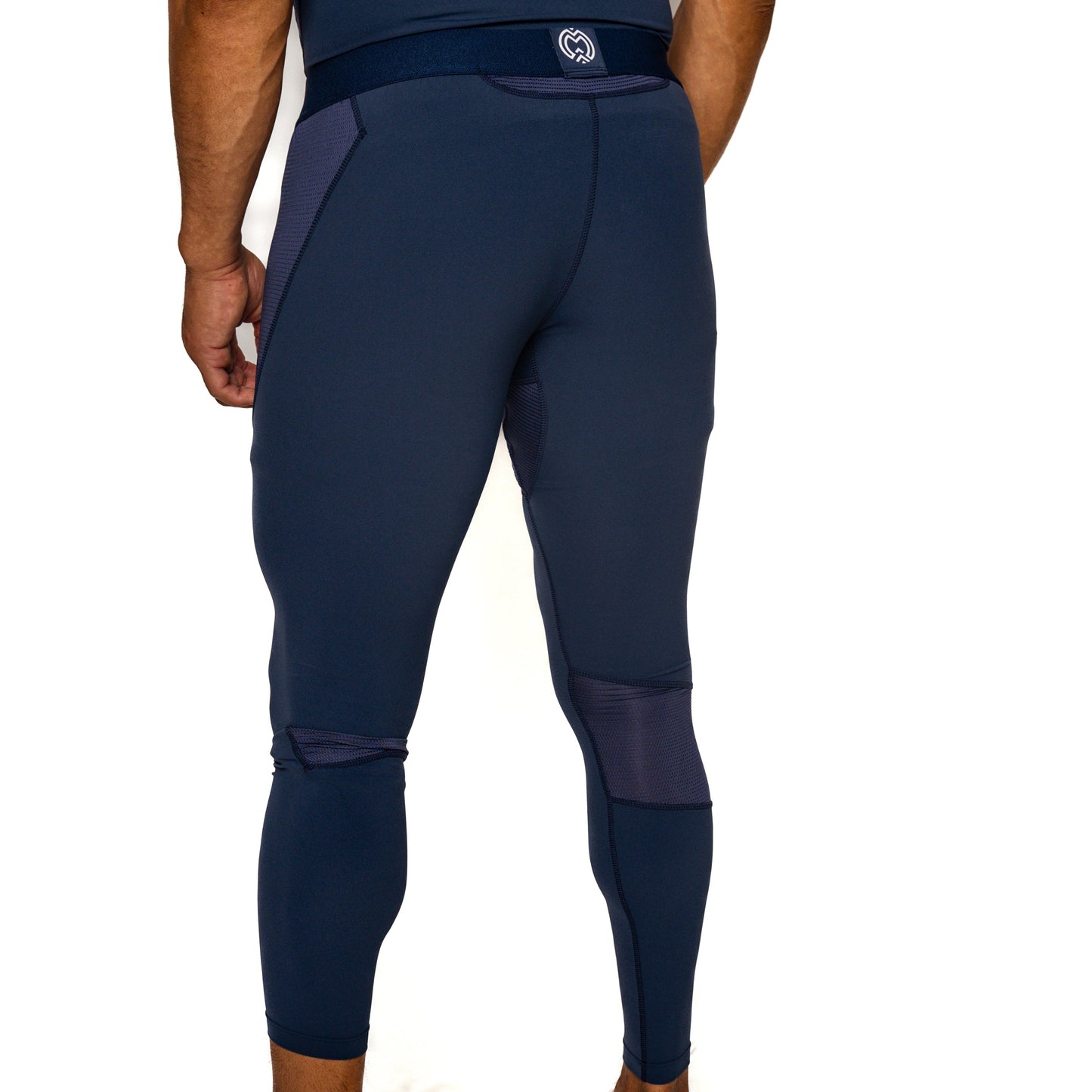 A boy faces away, wearing Massov ProForm® Boys' Compression Pants in navy blue. These ankle-length tights feature mesh panels, a small white back waistband logo, stretchy compression fabric, and enhance leg muscle definition.