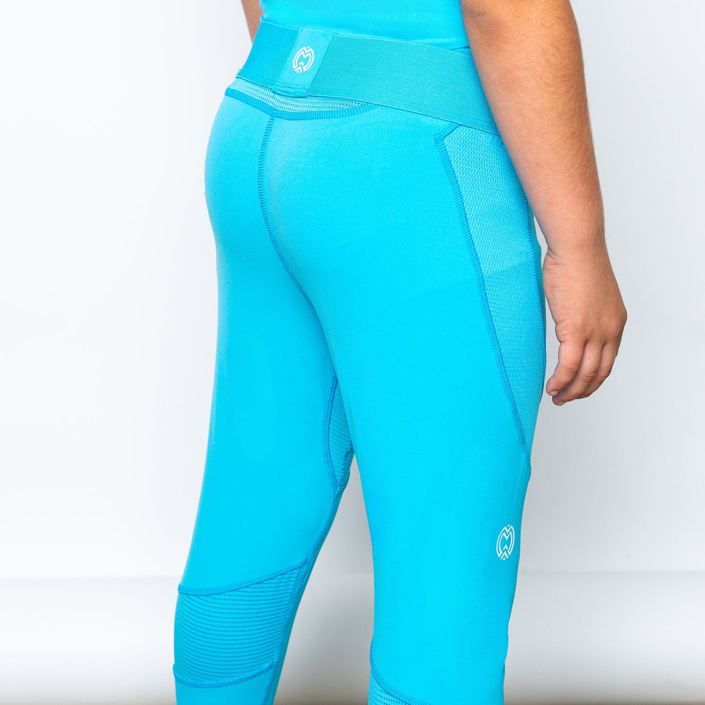 A boy faces away, highlighting the back view of Massov ProForm® Boys' Compression Pants—bright blue, with mesh accents and a white logo on the waistband and lower leg. The snug compression pants stand out against a plain white background.