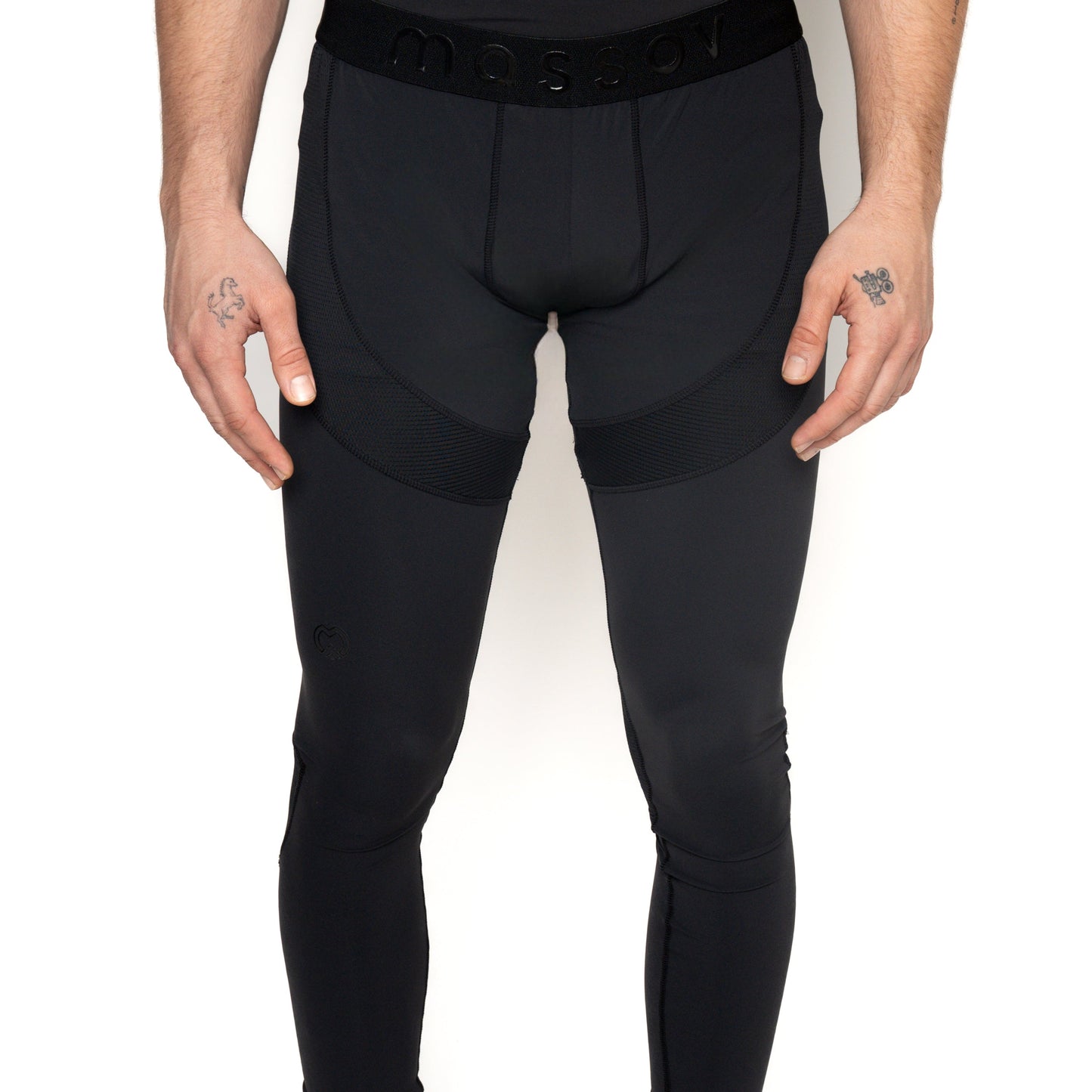 A person wears black Massov ProForm® Boys' Compression Pants with a visible MASSOV waistband. Their tattooed hands rest at their sides. The photo is cropped from mid-torso to feet against a plain white background.