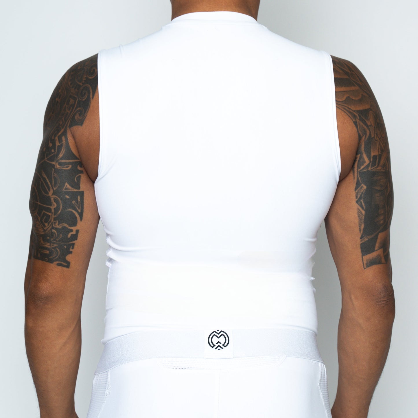 A person is shown from behind wearing the Massov ProForm® Boys' Compression Tank Top in white, which fits snugly to highlight tan skin and bold black arm tattoos. A small black Massov logo appears on the waistband of the matching bottoms.