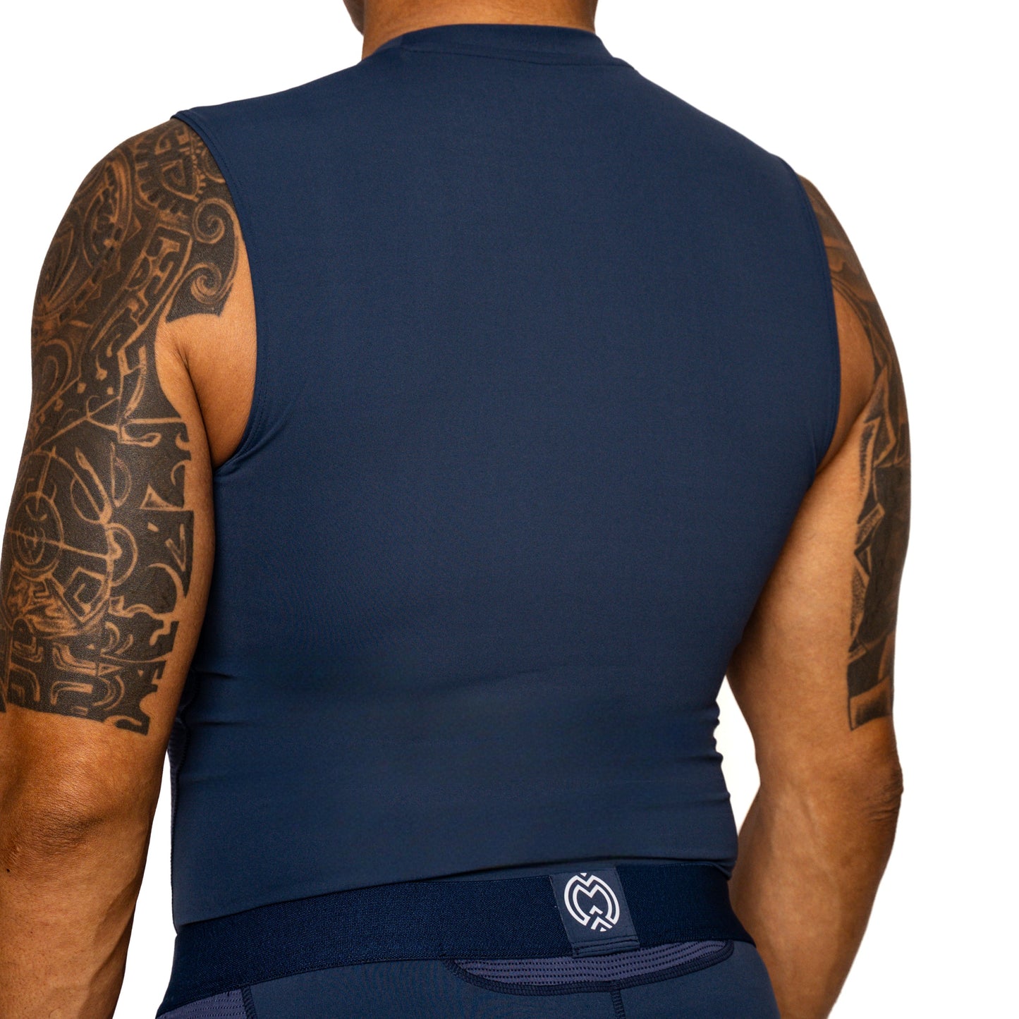 A person with tattooed arms is shown from the back wearing a Massov ProForm® Boys' Compression Tank Top in dark blue and matching shorts with a small white logo on the waistband, set against a plain white background.