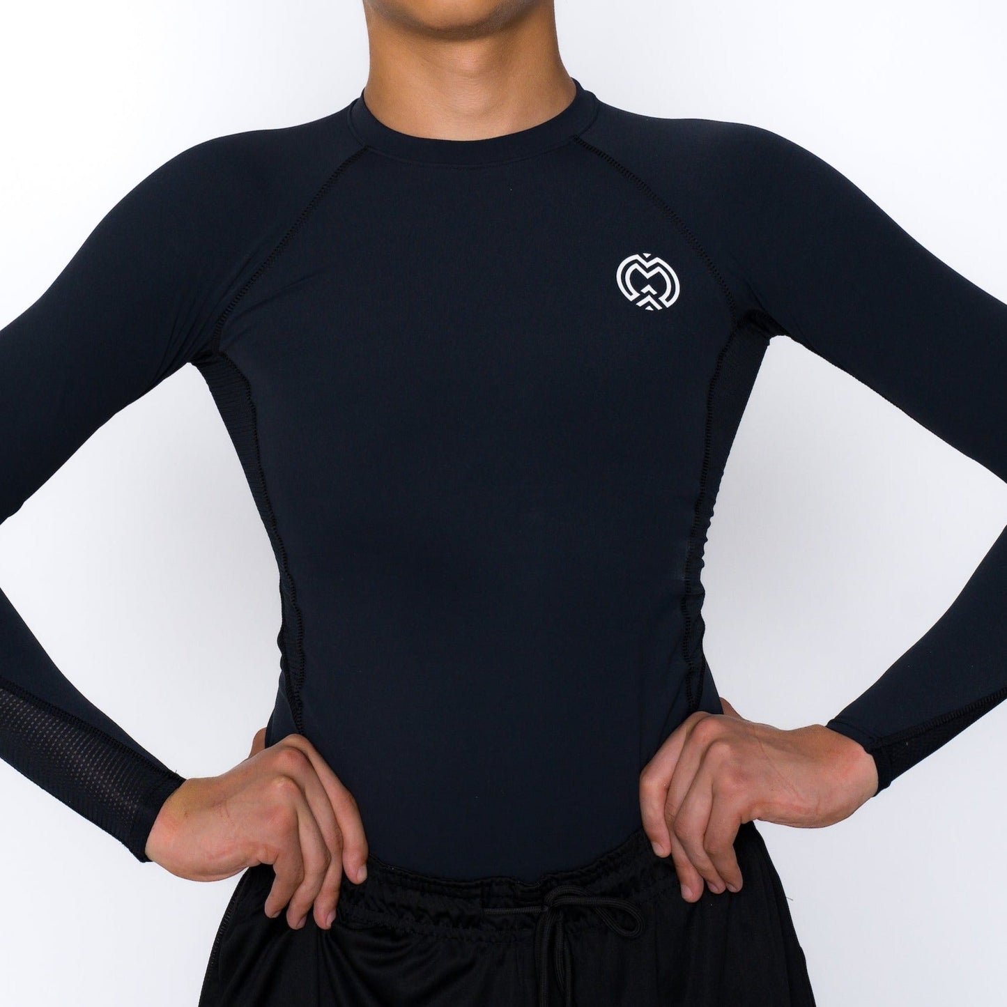 A person wearing the Massov ProForm® Boys' Long Sleeve Compression Shirt—fitted, black with a small white logo on the left chest—stands hands on hips against a plain background. The athletic shirt is paired with black pants; face not visible.