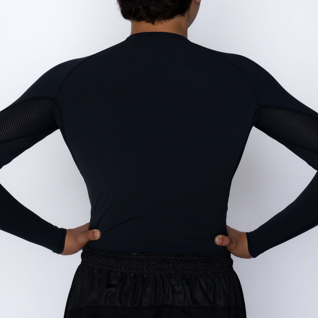 A youth boy stands with hands on hips, wearing a Massov ProForm® Boys' Long Sleeve Compression Shirt in black. The plain white background emphasizes his upper body and the matching black shorts.