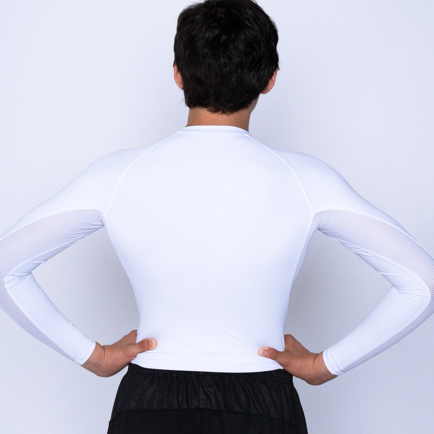 A youth boy with short dark hair stands with his back to the camera, hands on hips, wearing a fitted white ProForm® Boys' Long Sleeve Compression Shirt by Massov and black shorts. The background is plain and light-colored.