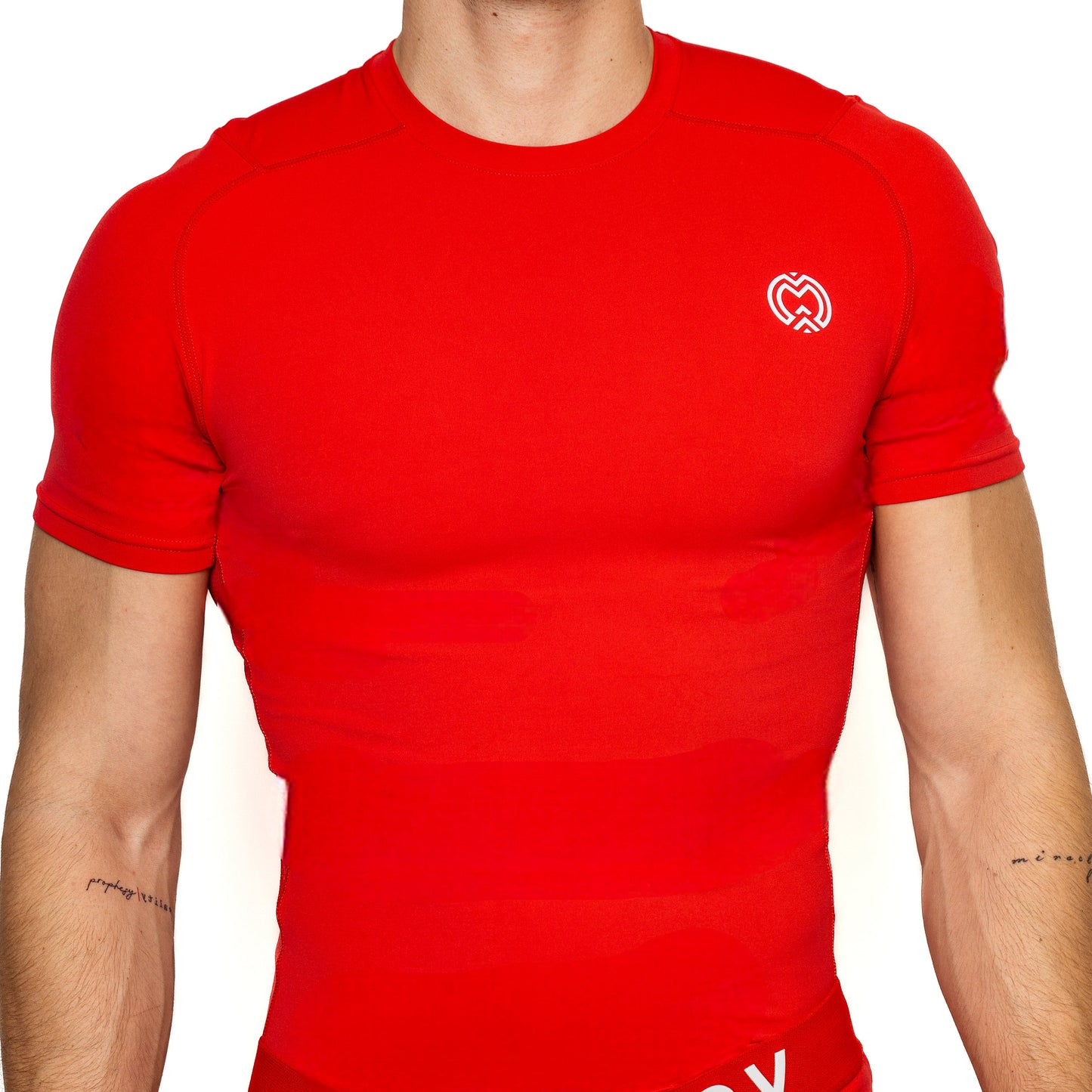 A person wearing the Massov ProForm® Boys' Short Sleeve Compression Shirt in bright red with UPF protection and a small white heart logo on the left chest. The image highlights their upper torso and tattooed forearms against a plain background.