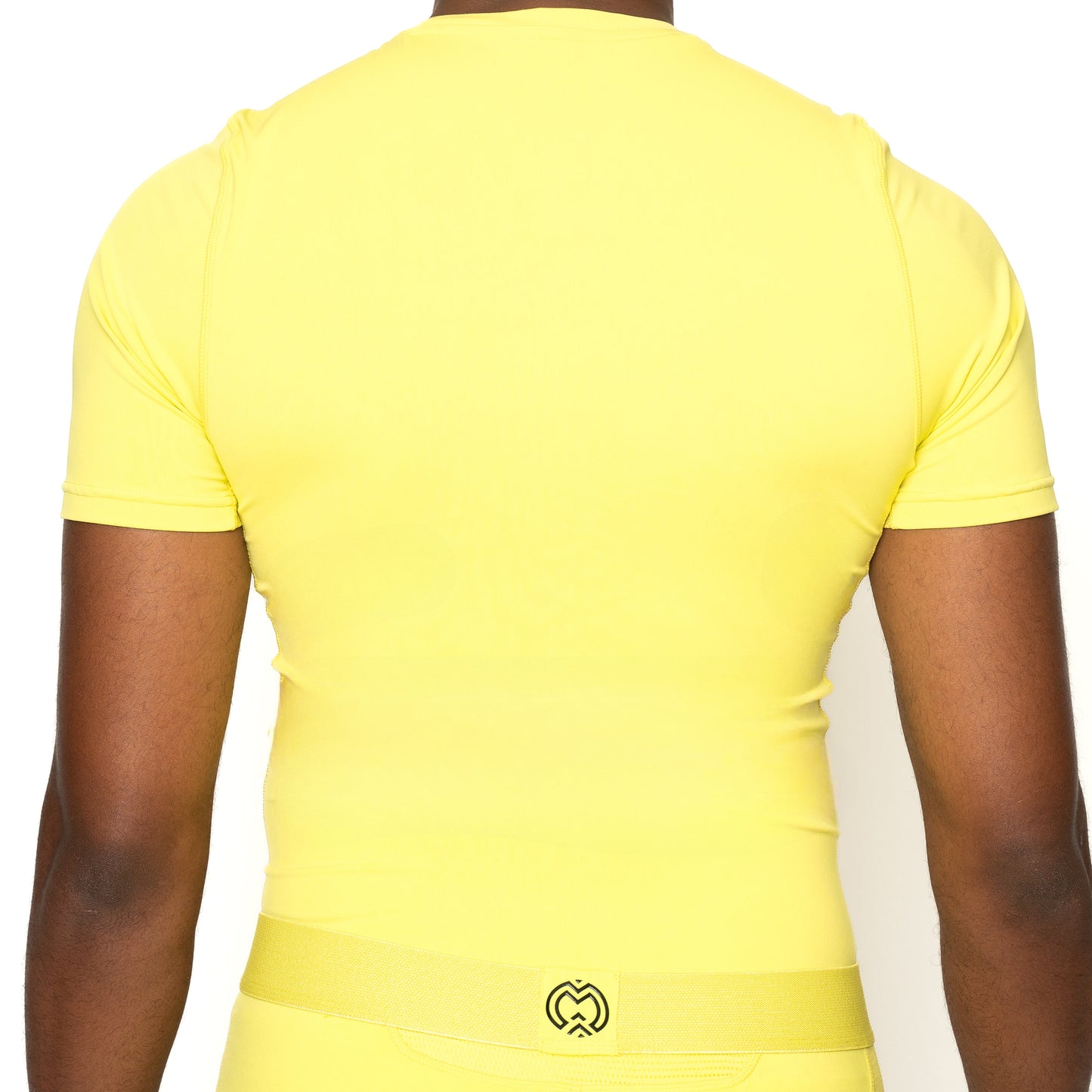 A boy models the Massov ProForm® Boys' Short Sleeve Compression Shirt in yellow, with matching pants. He faces away, arms at his sides, showing a small black heart-shaped logo on the waistband against a white background.