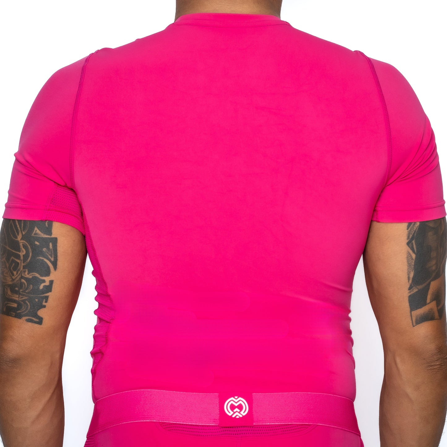 A tattooed person is shown from behind in a Massov ProForm® Boys' Short Sleeve Compression Shirt and matching waistband, both bright pink with a white heart-shaped logo on the waistband, set against a plain white background.