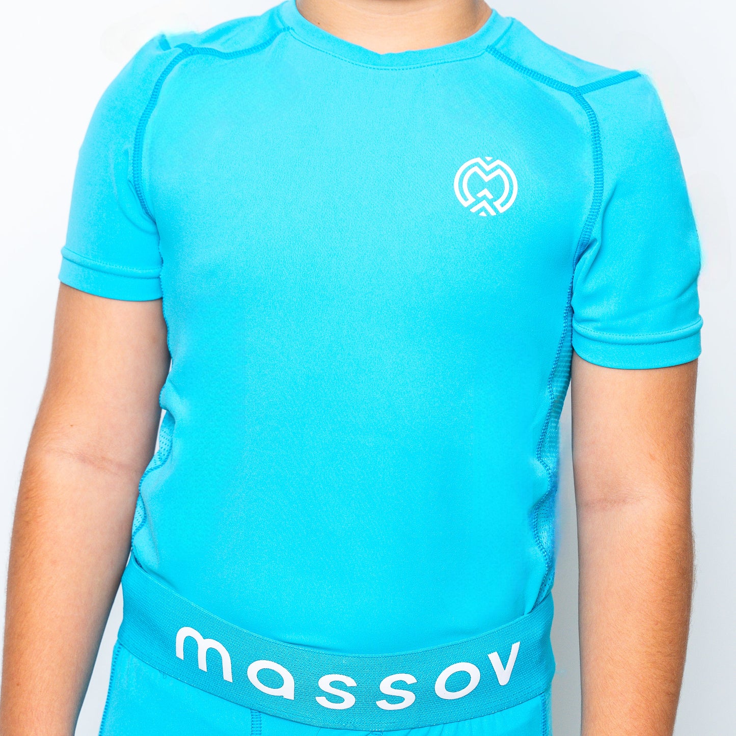 A child wears a bright blue ProForm® Boys' Short Sleeve Compression Shirt by Massov with a white left-chest logo, paired with matching shorts featuring a blue waistband and "massov" in white. The background is plain and light-colored.