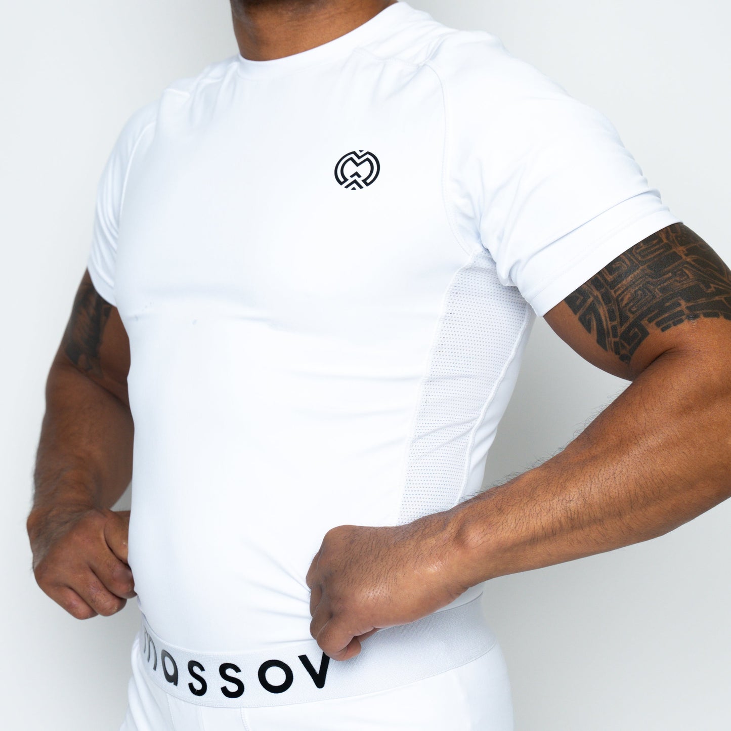A person in a tight Massov ProForm® Boys' Short Sleeve Compression Shirt with a small black logo stands against a white background. Their tattooed forearms are visible, and the athletic shirt provides UPF protection for outdoor activities.