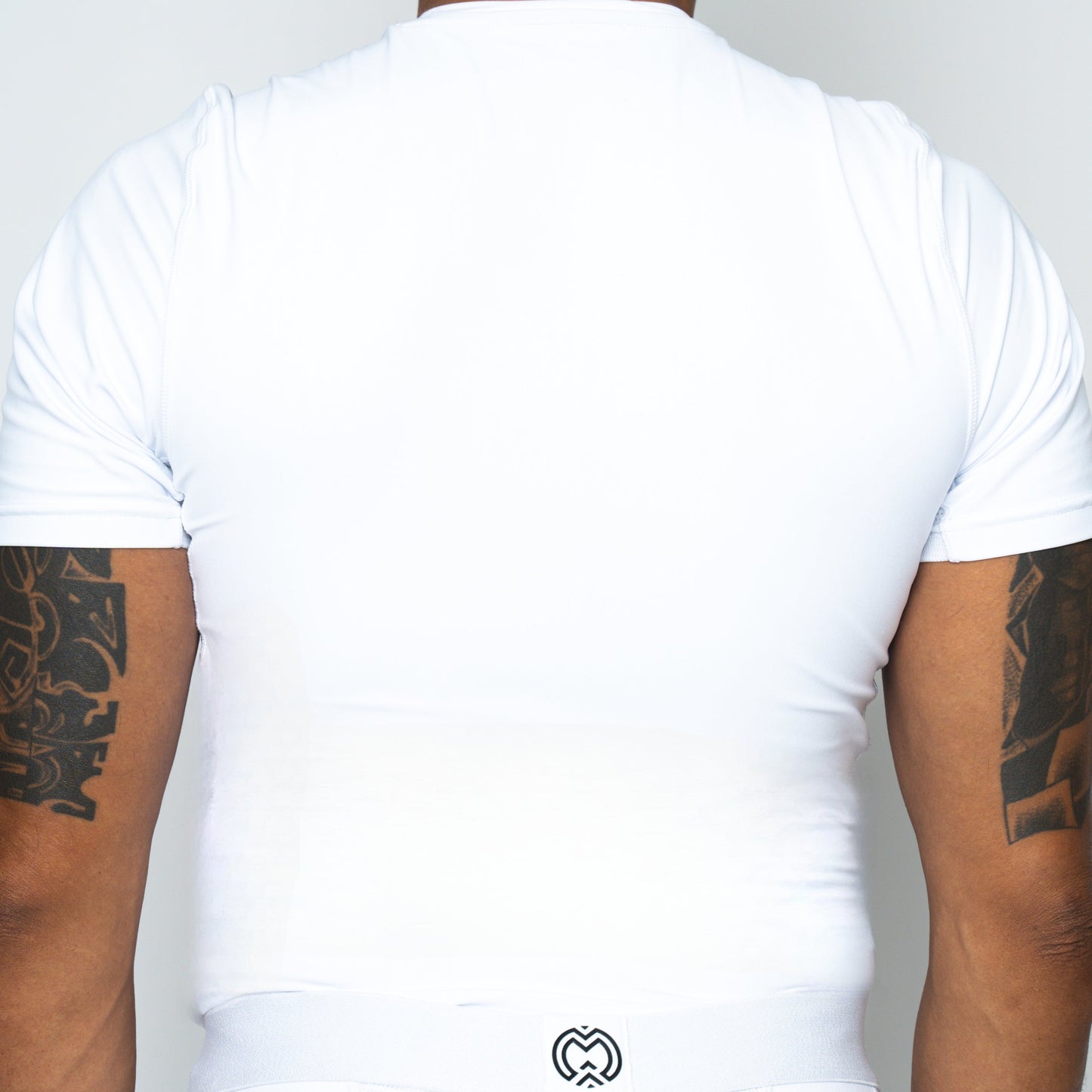 A person is seen from the back wearing a Massov ProForm® Boys' Short Sleeve Compression Shirt. The fitted white design and small black logo at the bottom stand out against a light background, while dark, intricate tattoos cover both arms.