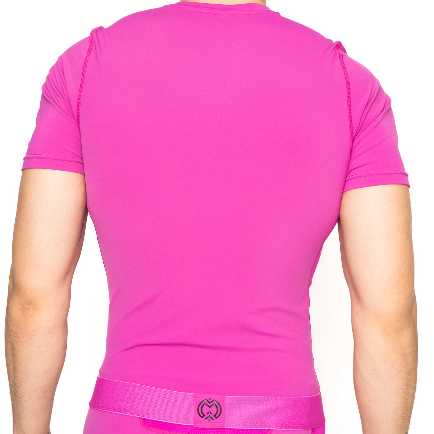 A boy faces away from the camera wearing a vibrant pink Massov ProForm® Boys' Short Sleeve Compression Shirt and matching waistband, with his back, arms, and waist visible against a plain white background.
