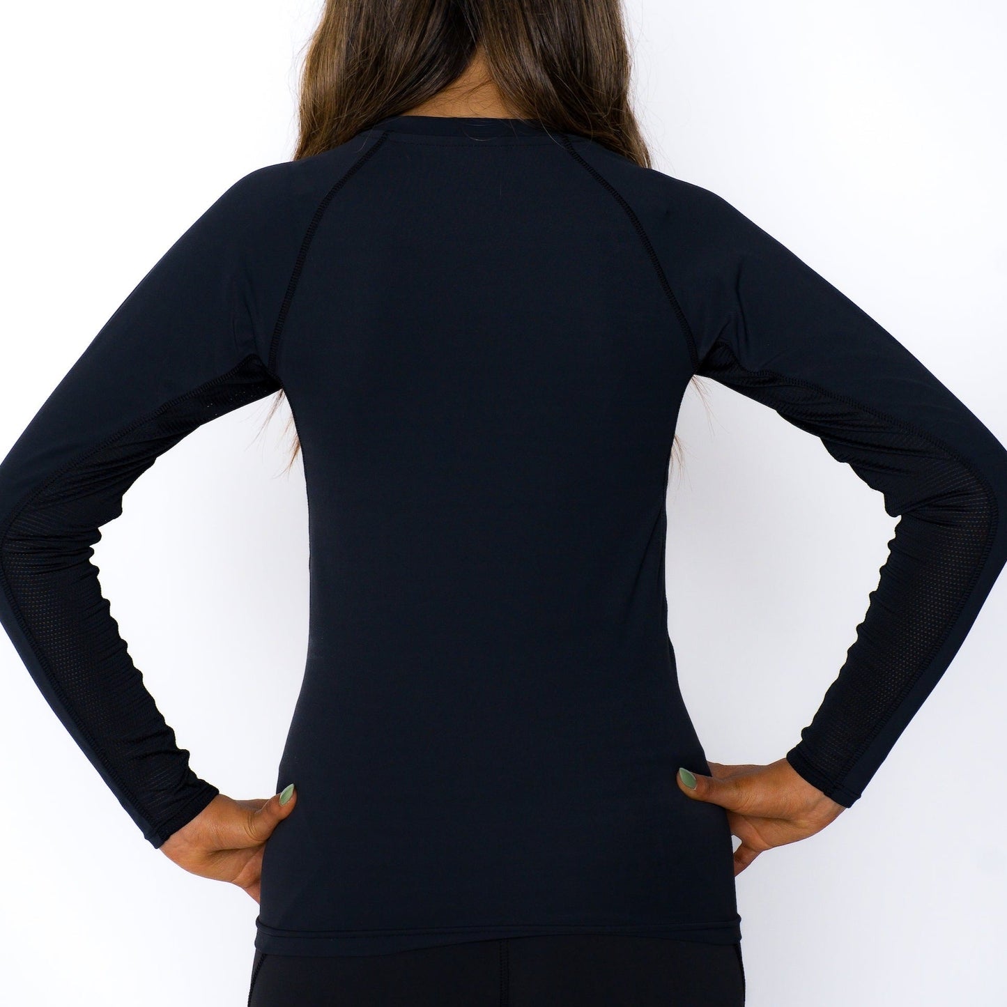 A girl with long brown hair, facing away from the camera, wears the Massov ProForm® Girls' Long Sleeve Compression Shirt. Her hands are on her hips with elbows bent, set against a plain white background.